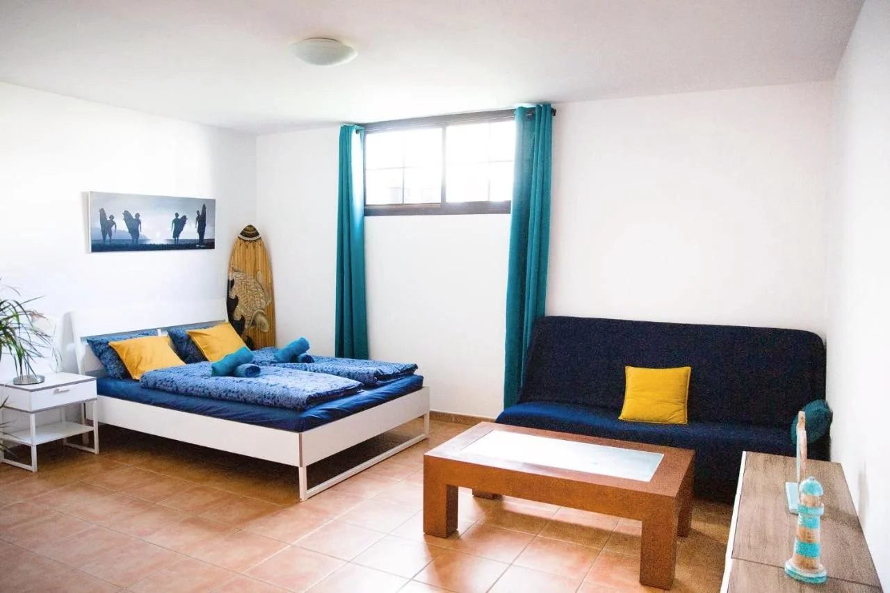 Dorm room with a white bed, blue bedding, yellow pillows, and a small white nightstand with a plant. There is a black sofa with a yellow pillow, a wooden coffee table, and a window with blue curtains. An surfboard is standing next to the bed and a piece of art is on the wall.