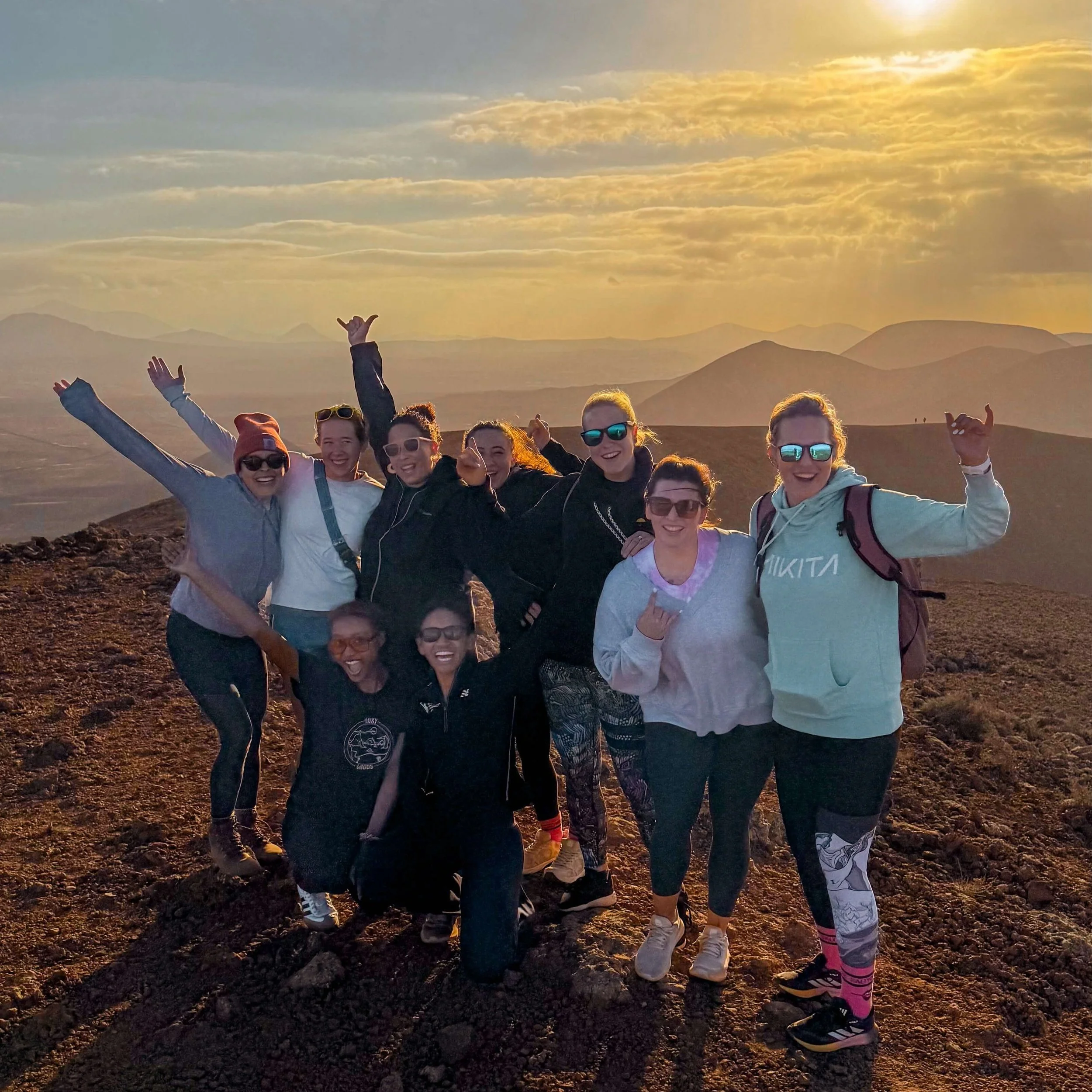 Group of eight women hiking on a mountain at sunset, smiling and making various hand gestures.