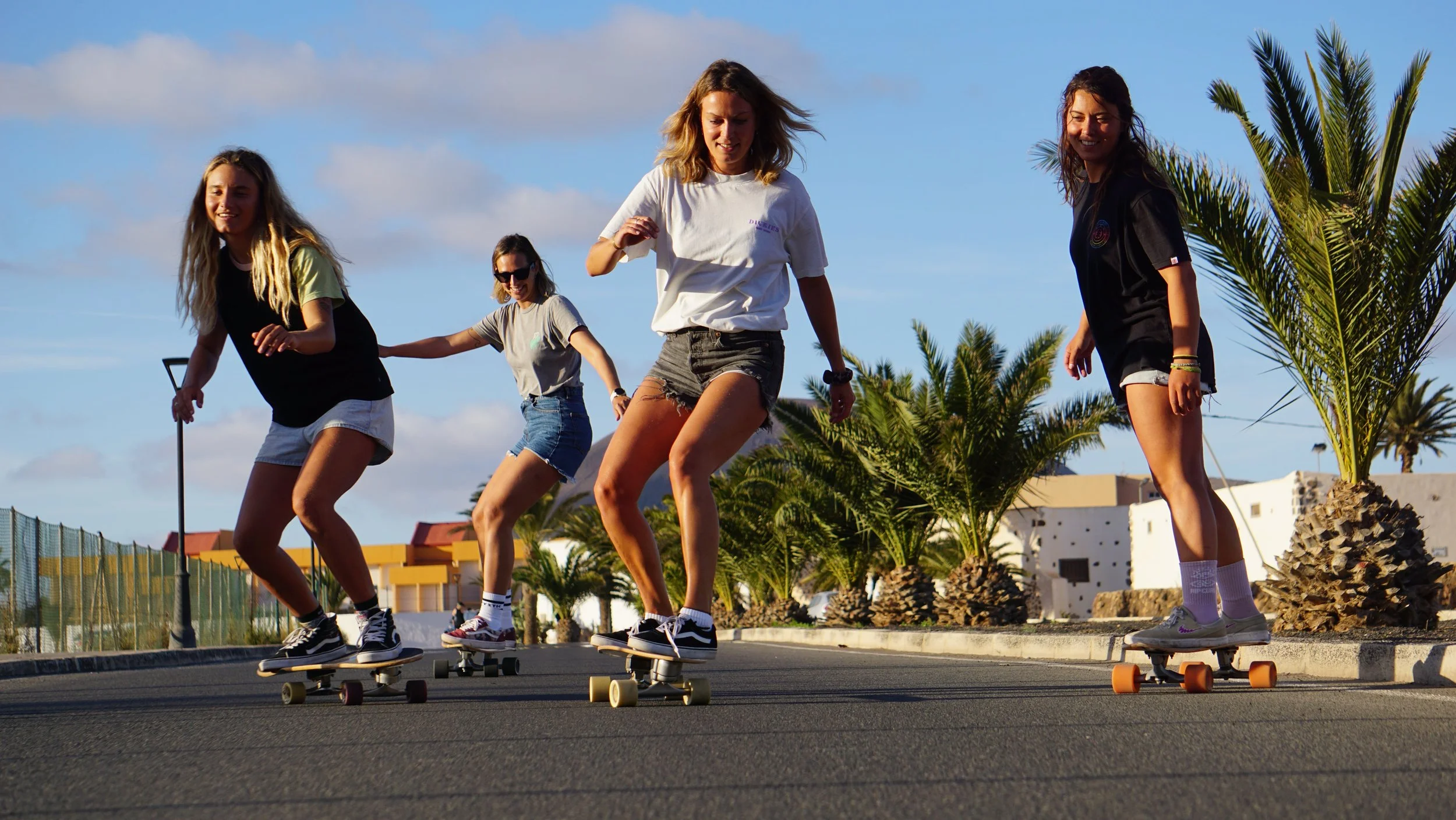 Four young women skateboarding on a street with palm trees and buildings in the background