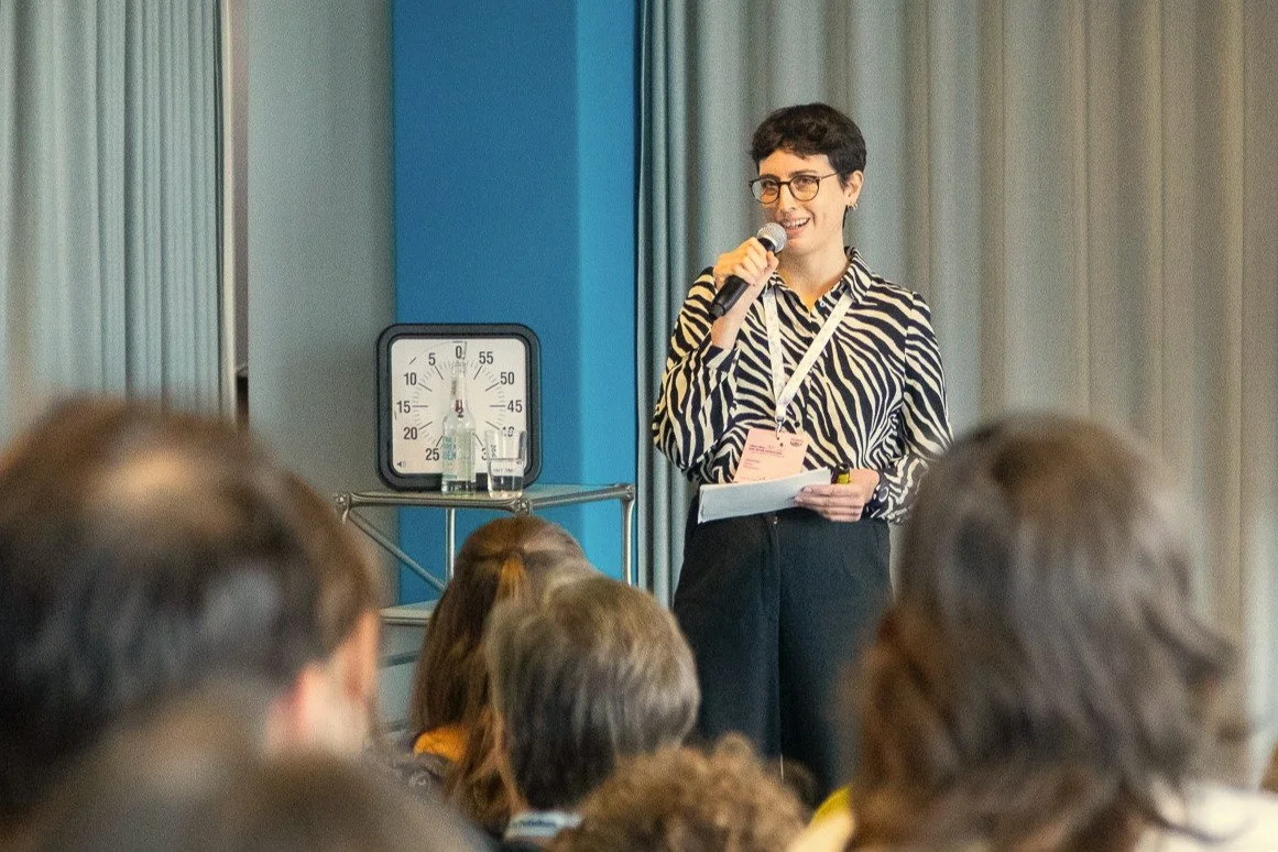 Eine Frau mit kurzen dunklen Haaren und Brille spricht bei einer Veranstaltung vor Publikum, im Hintergrund steht ein Timer.