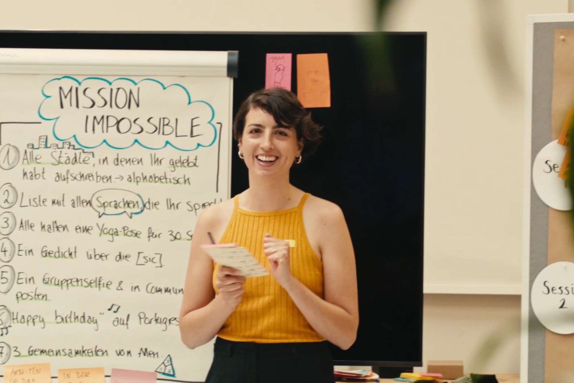 Eine lächelnde Frau in einem gelben ärmellosen Oberteil hält einen Notizblock und einen Stift in den Händen, vor einem Whiteboard mit dem Schriftzug 'Mission Impossible' und einer Liste. Sie steht in einem Raum mit Post-its und Pinnwänden.