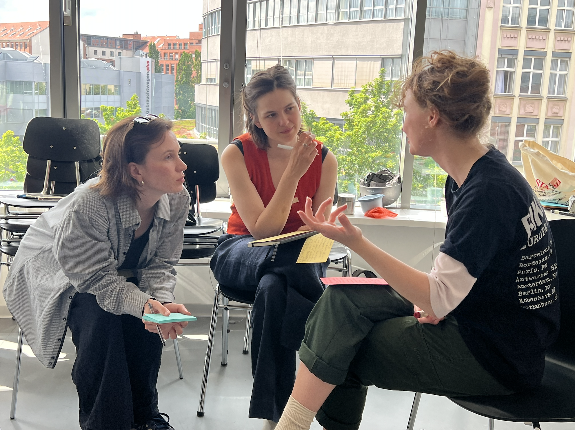 Drei Frauen diskutieren in einem Raum mit großen Fenstern, durch die bunte Stadtarchitektur zu sehen ist.