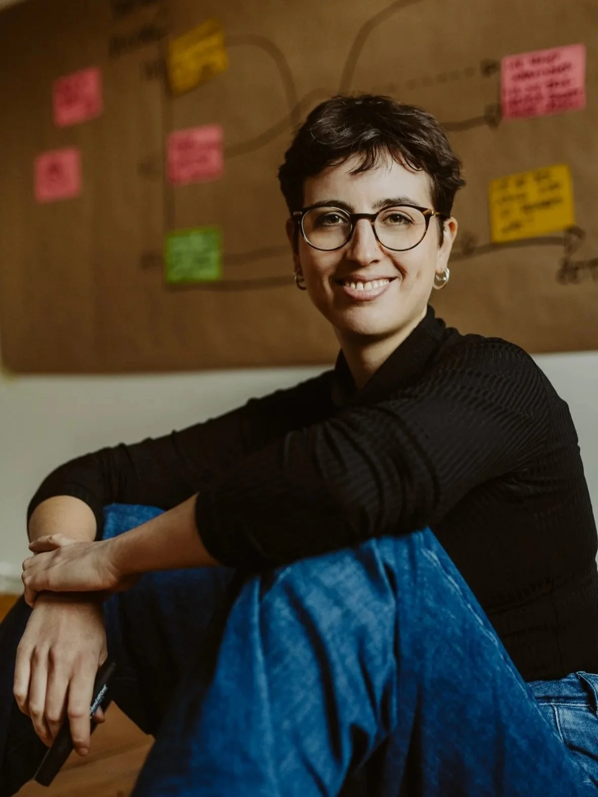 Lächelnde Frau mit kurzen dunklen Haaren und Brille sitzt locker vor einer Wand mit bunten Post-its