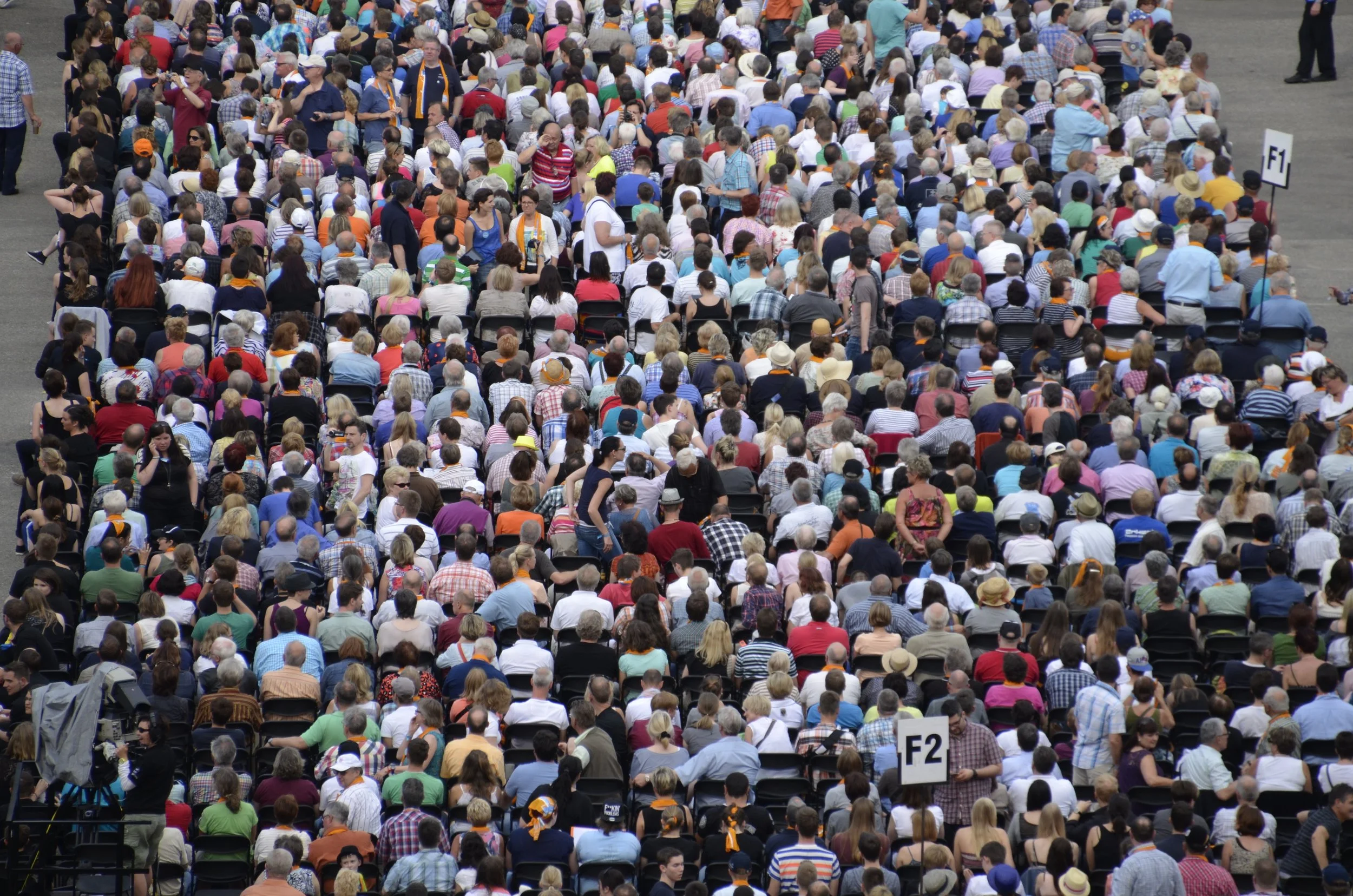 Une grande foule de personnes assises en rangées, lors d'une cérémonie ou d'un événement public, avec des panneaux indiquant F1 et F2.