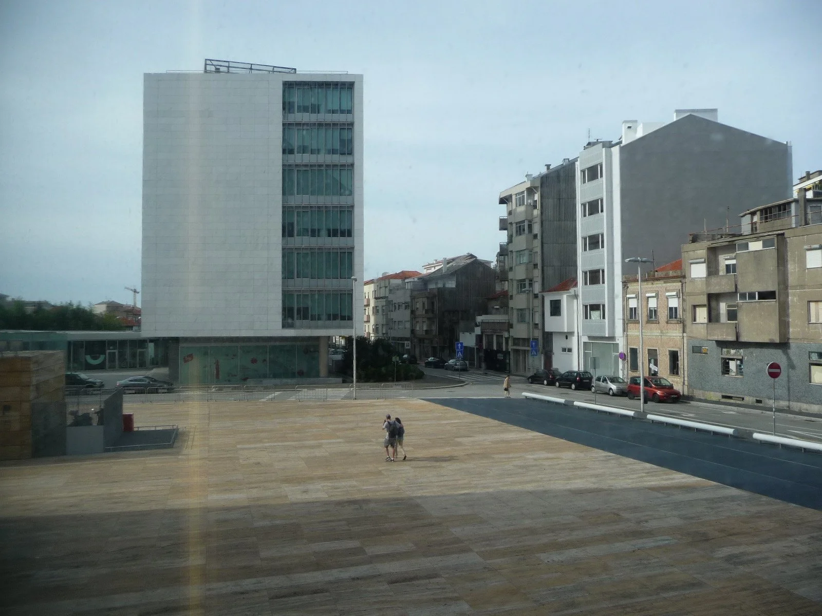 Vue de plusieurs immeubles résidentiels et commerciaux, avec une grande place pavée au premier plan et deux personnes debout près du centre, en journée avec ciel nuageux.