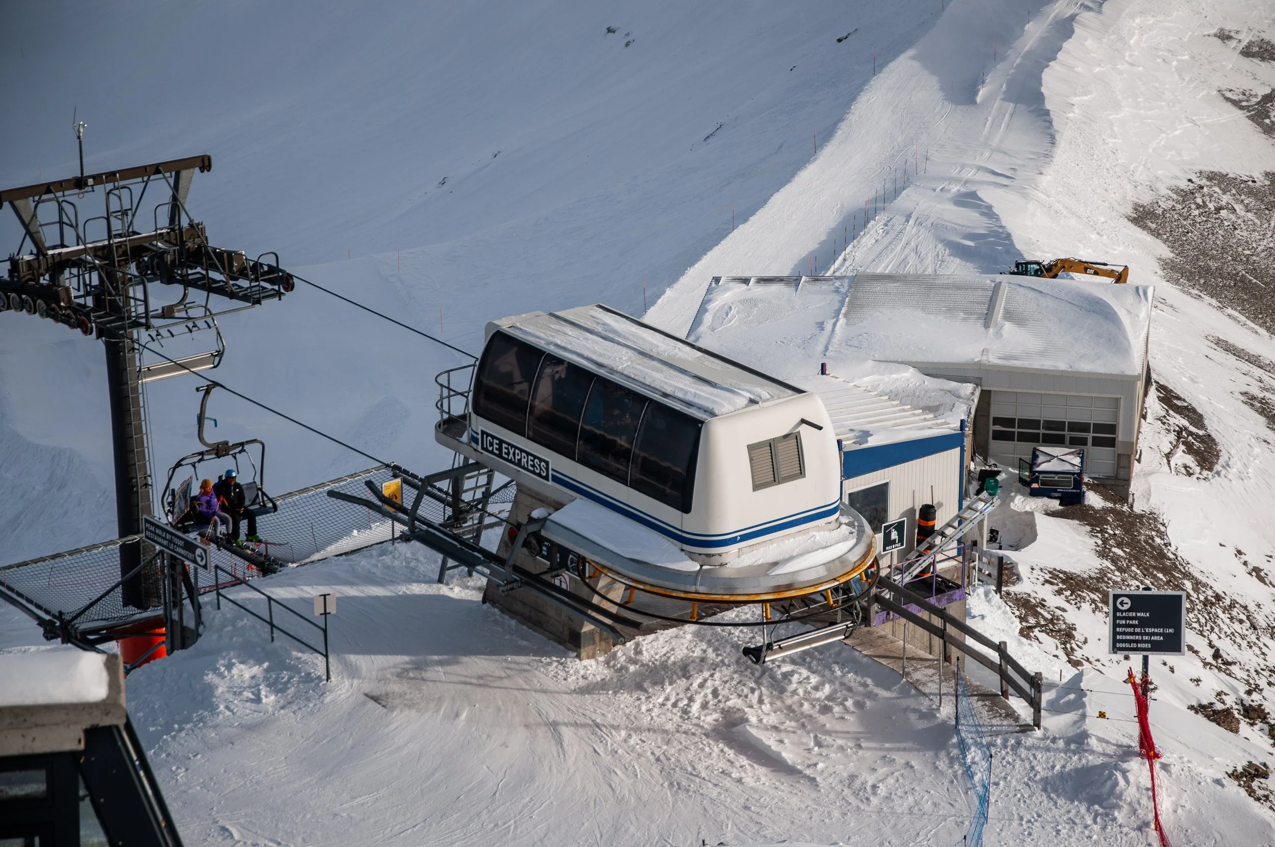 Un télésiège en station de ski avec deux personnes assises, un bâtiment pour la cabine du téléphérique, et un paysage enneigé de montagnes.