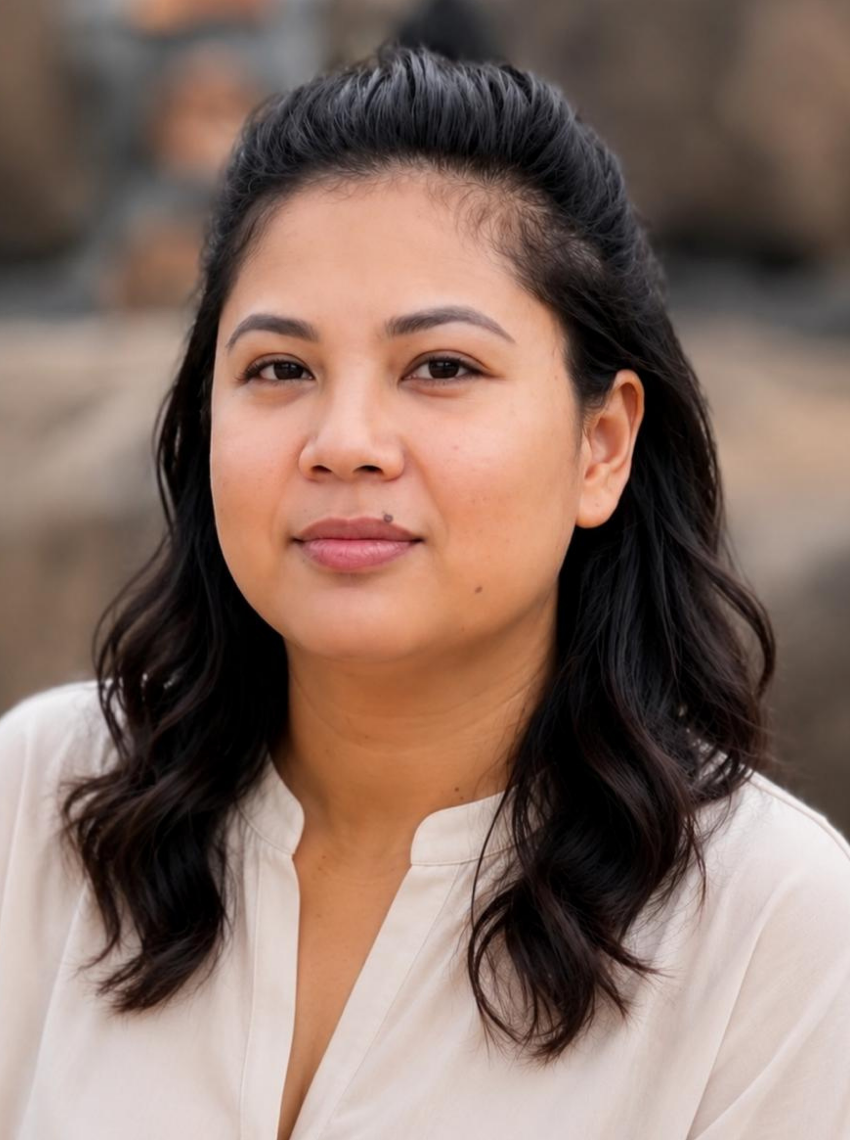 A close-up of a woman with dark, wavy hair, wearing a light-colored V-neck blouse, outdoors with blurred natural background.