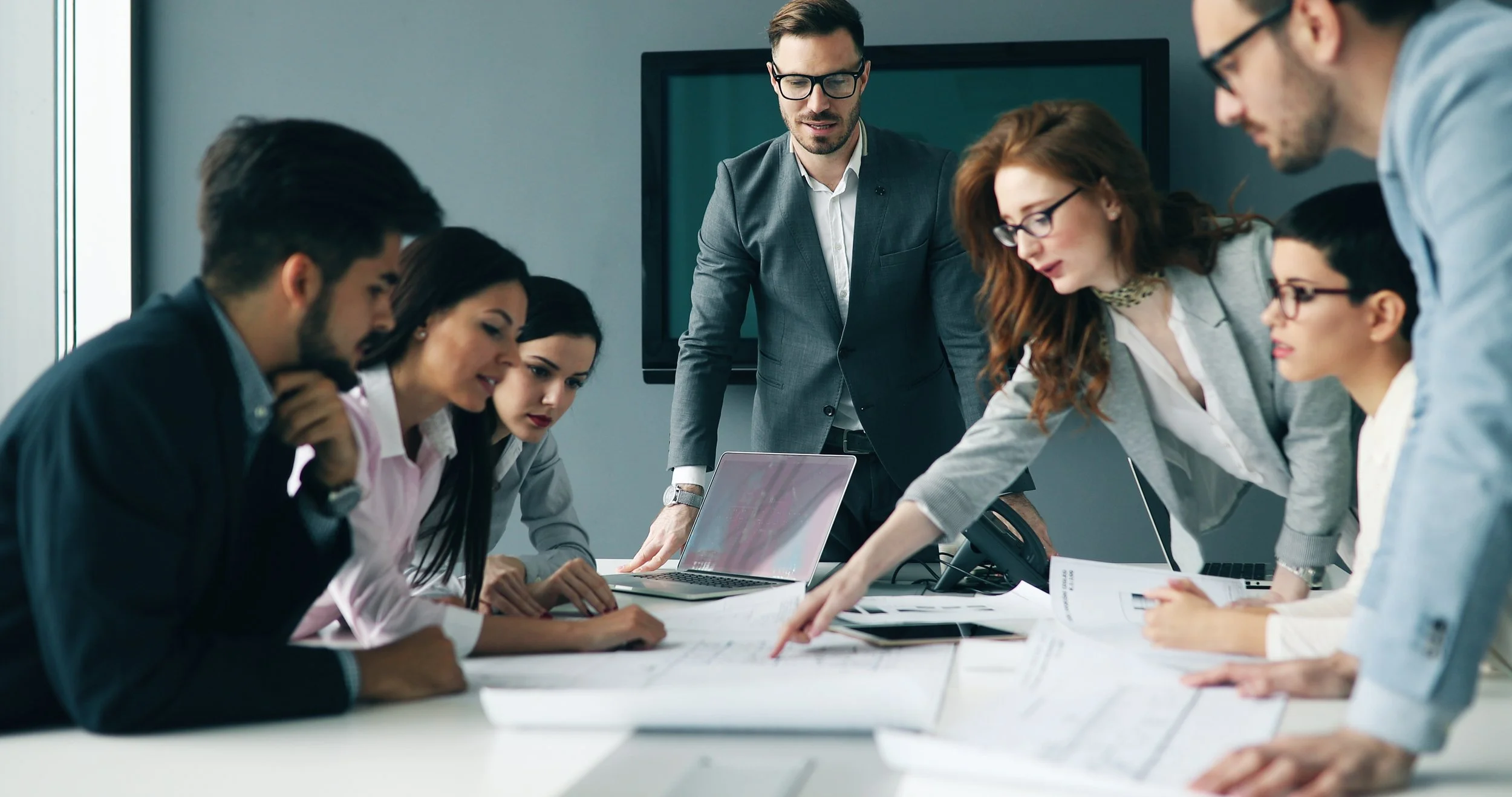 A picture of a team collaborating around a conference table reviewing documents and a laptop during a business meeting.