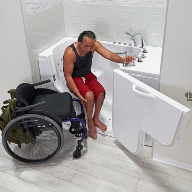 A woman in a wheelchair using a sink with an accessible door and faucet in a bathroom, with a bathtub nearby.