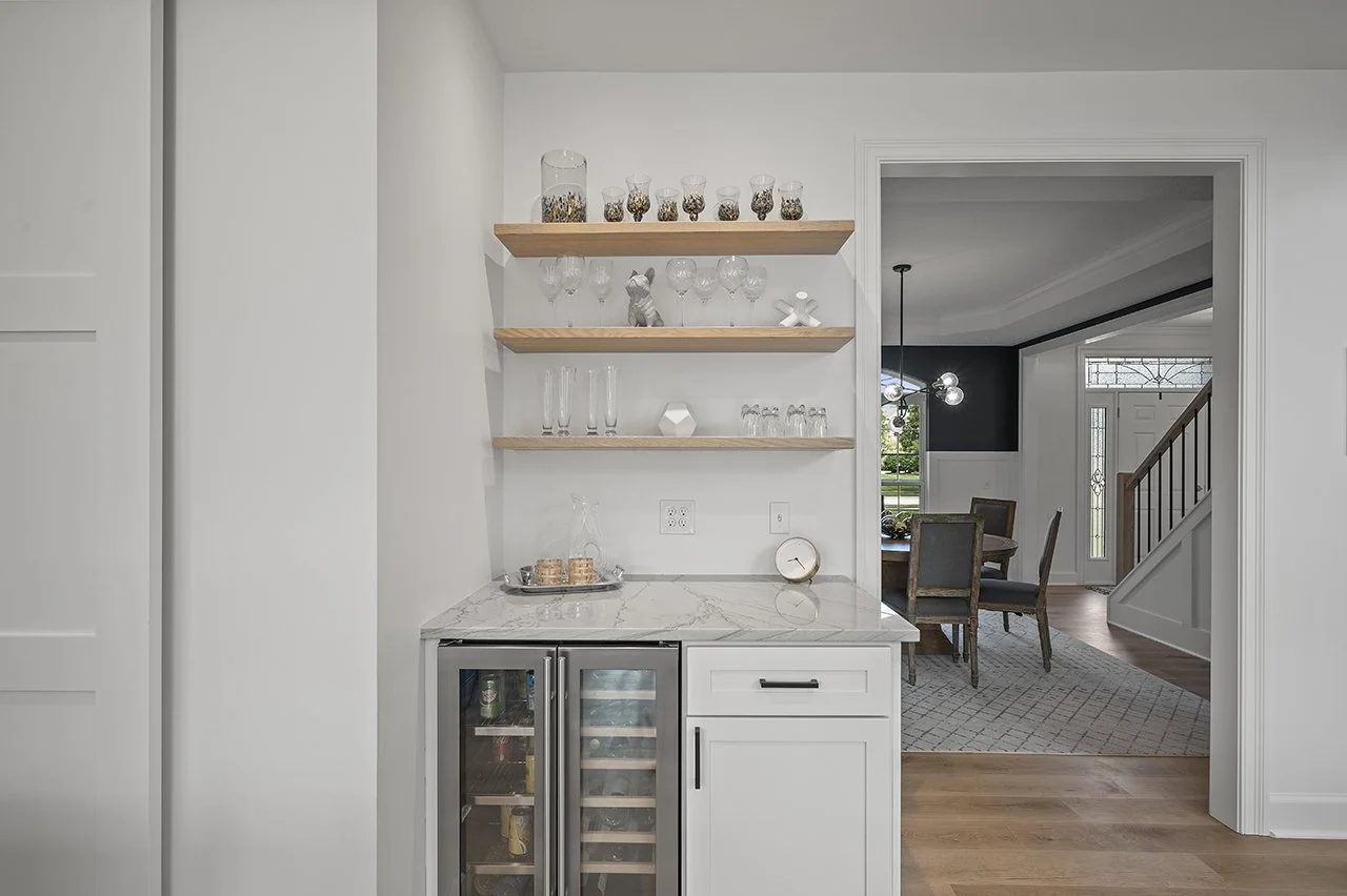 A bar area next to the pantry with a matching solid surface countertop. A beverage cooler with a glass front is next to a cabinet and open shelving is mounted to the wall.
