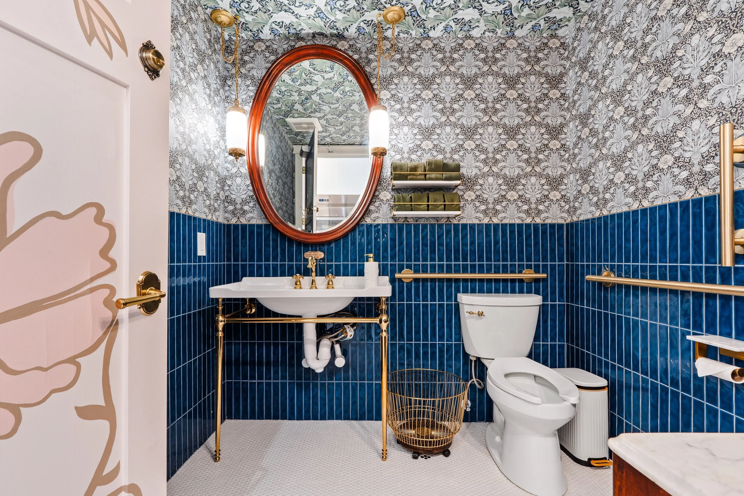 Bathroom with a half wall of blue tile and vintage inspired wallpaper.