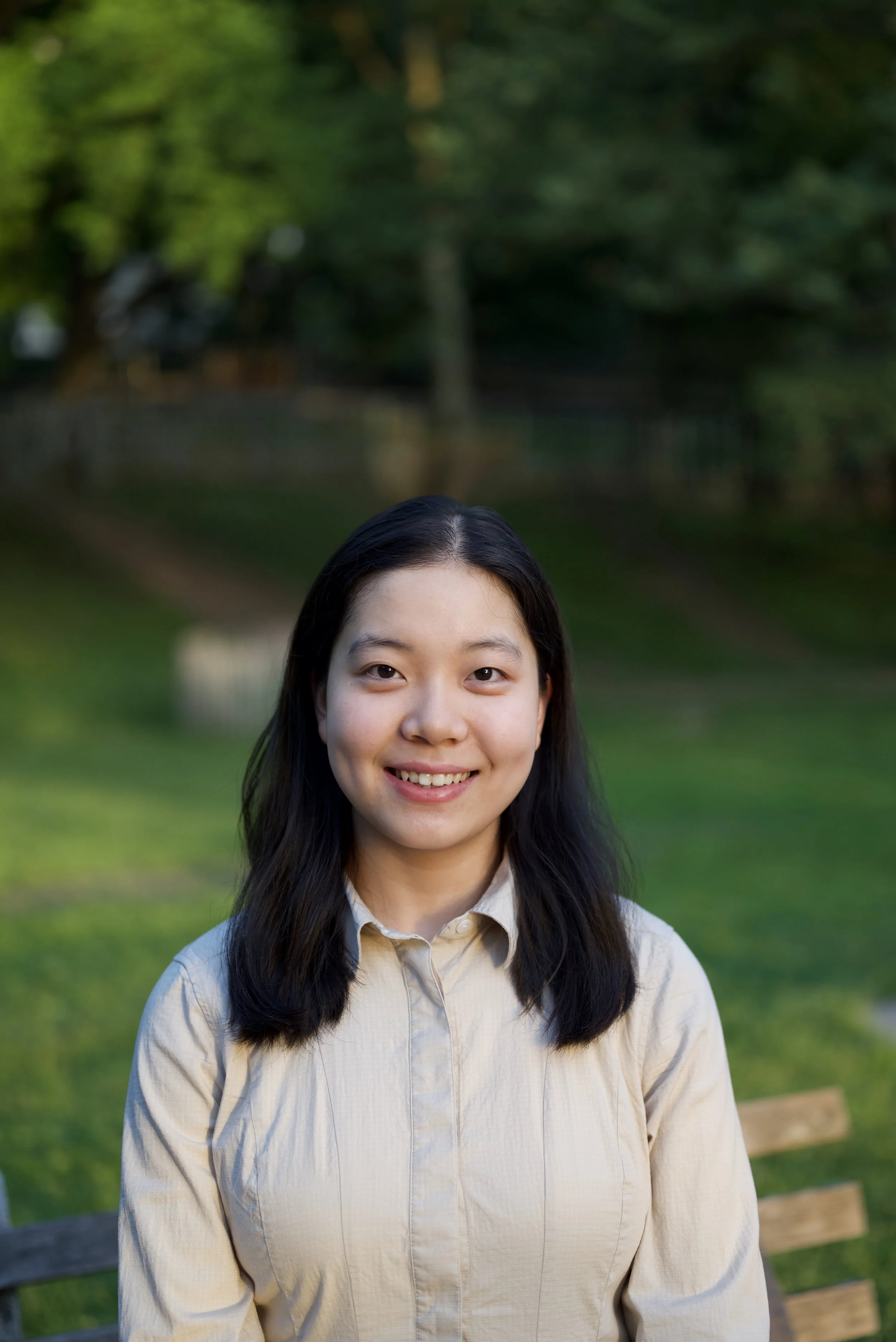 A young woman with long black hair standing outdoors in front of a green, wooded background, smiling at the camera.