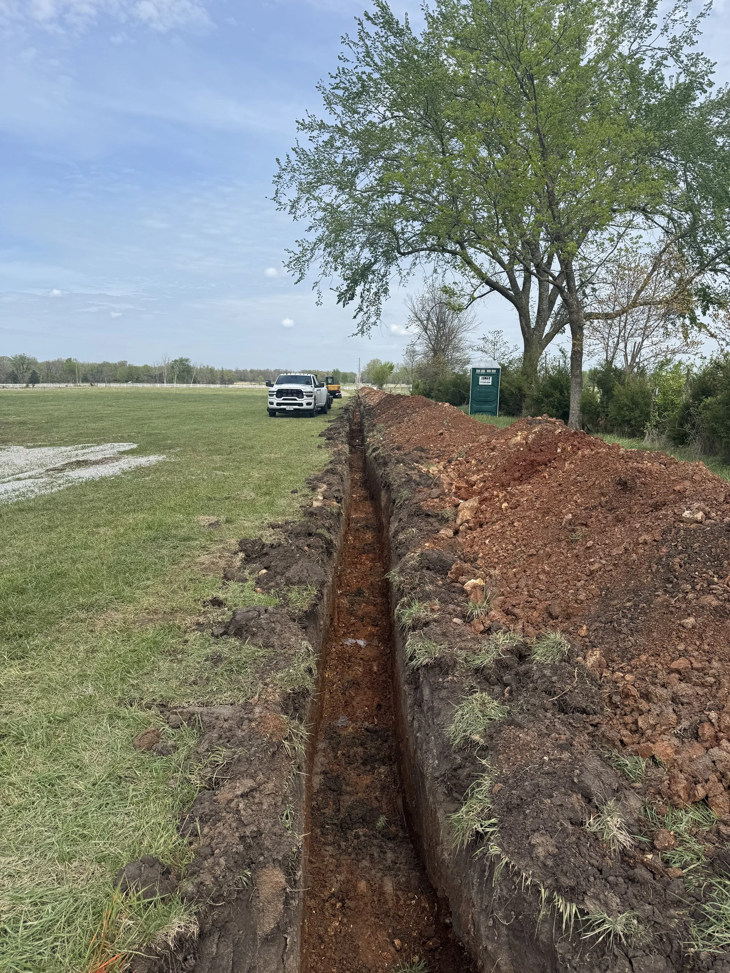 A construction site in a grassy field with a large tree, a portable toilet, and a trench being dug for utilities or drainage, with vehicles parked nearby.