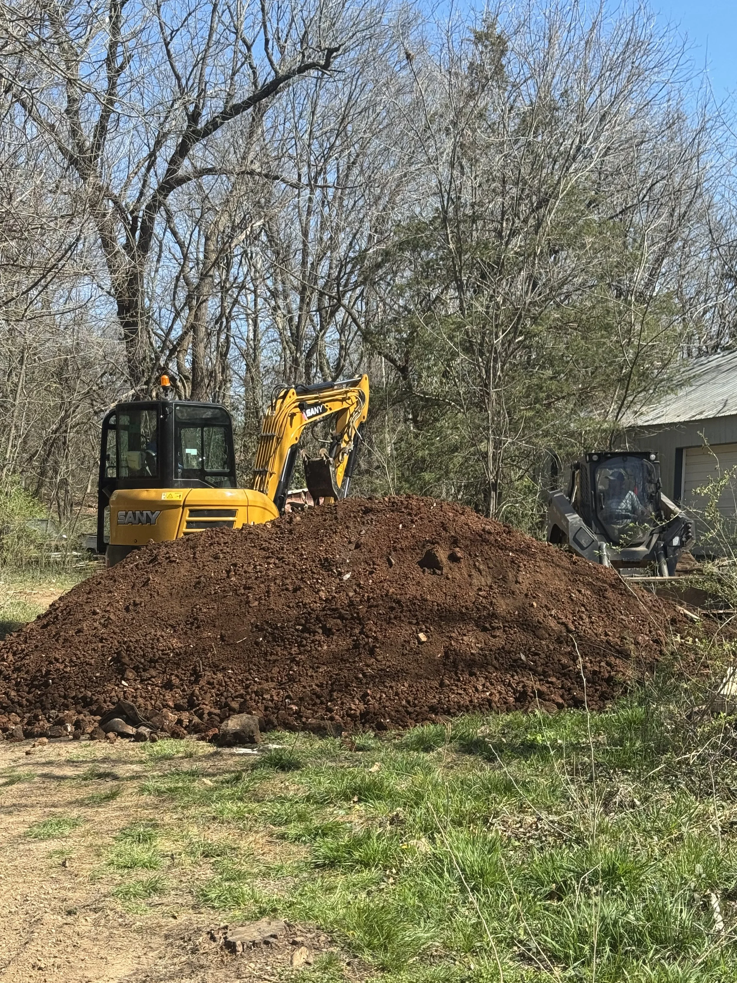 Construction site with a large pile of dirt, two small excavators, trees, and a building with a metal roof in the background.