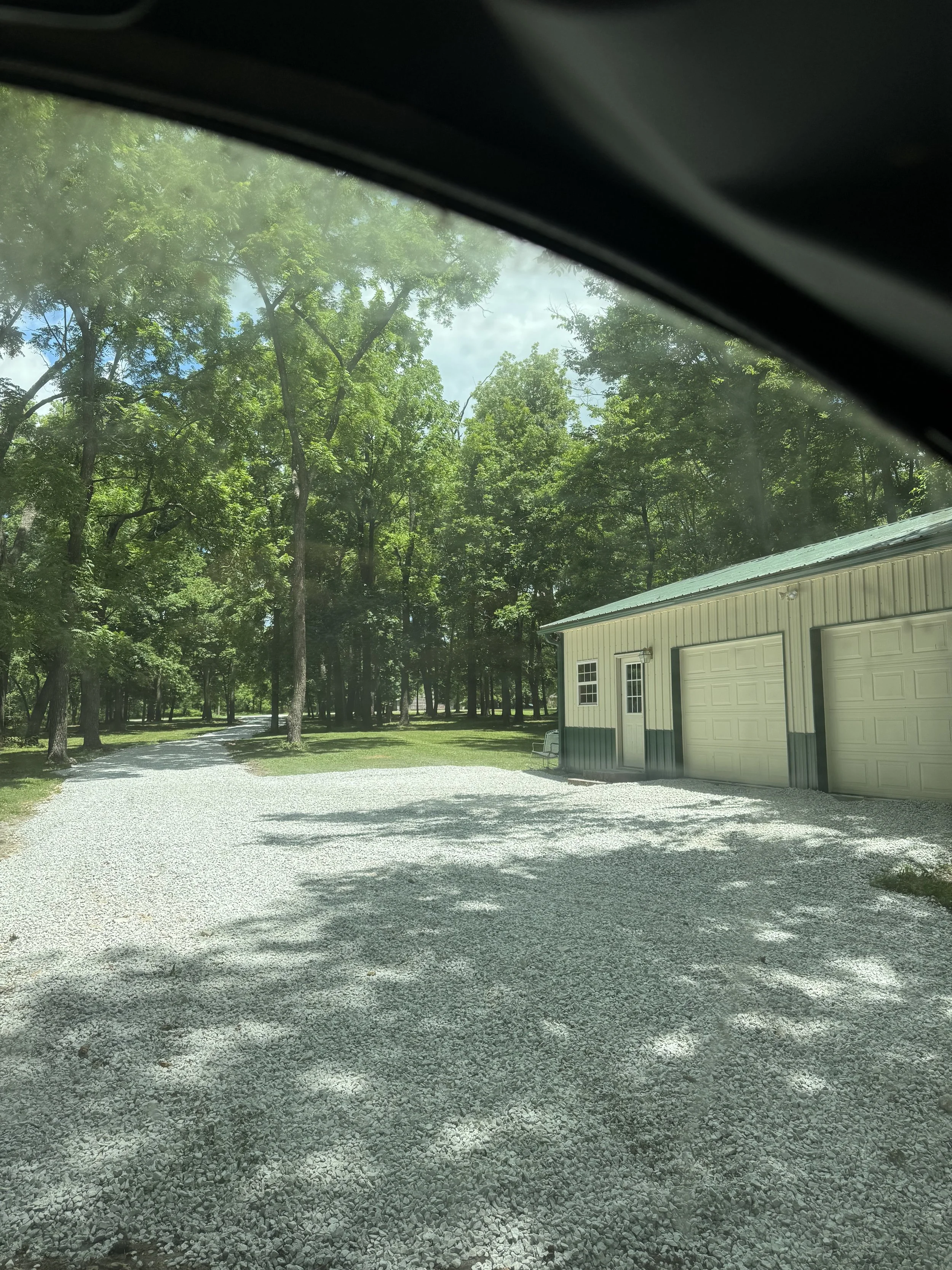 View of a gravel driveway leading to a garage with two doors, surrounded by green trees and shade.