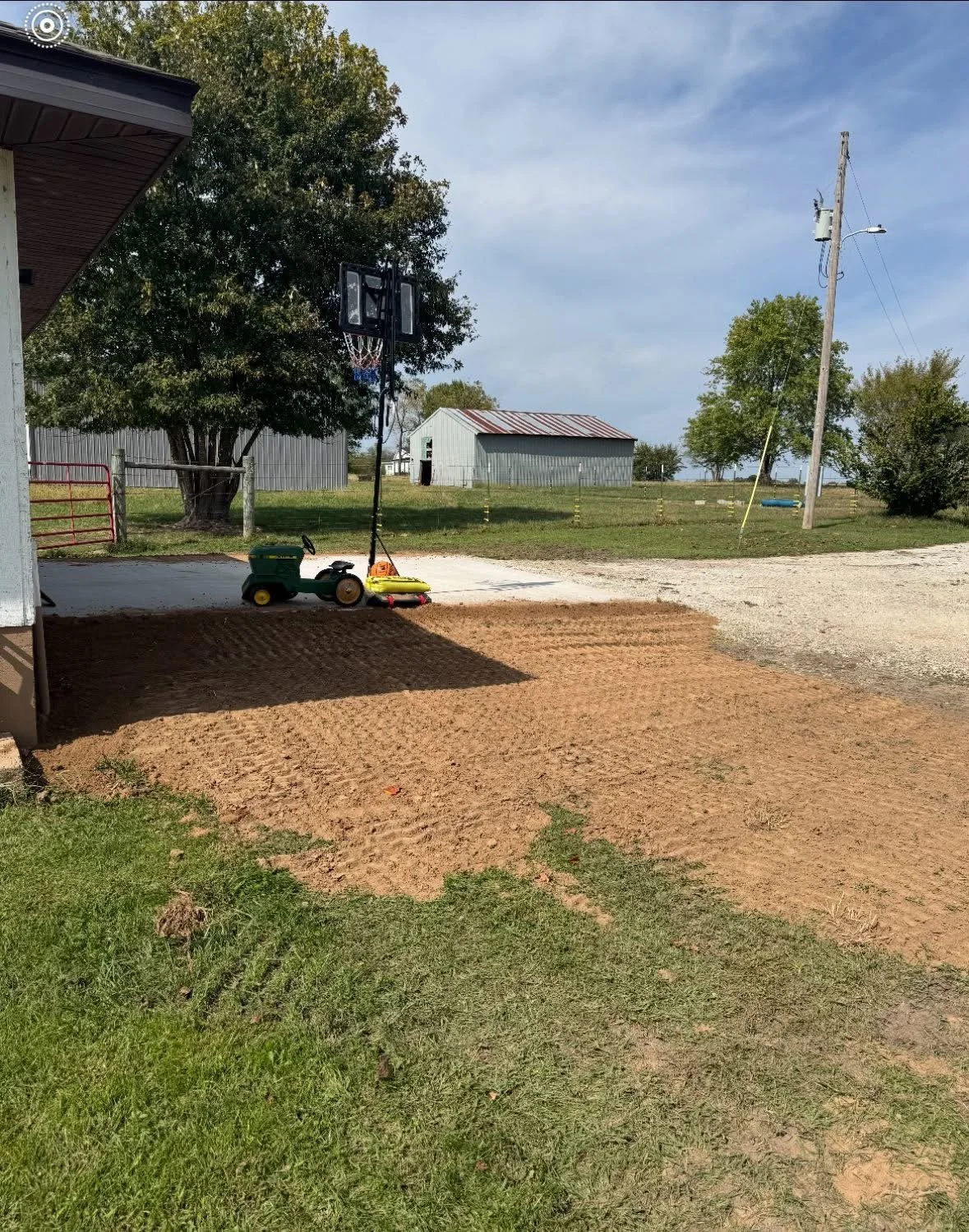 A backyard scene with a dirt patch, a small green riding lawnmower, a yellow and red toy lawnmower, a basketball hoop, a large tree, a metal shed, and a utility pole, under a partly cloudy sky.