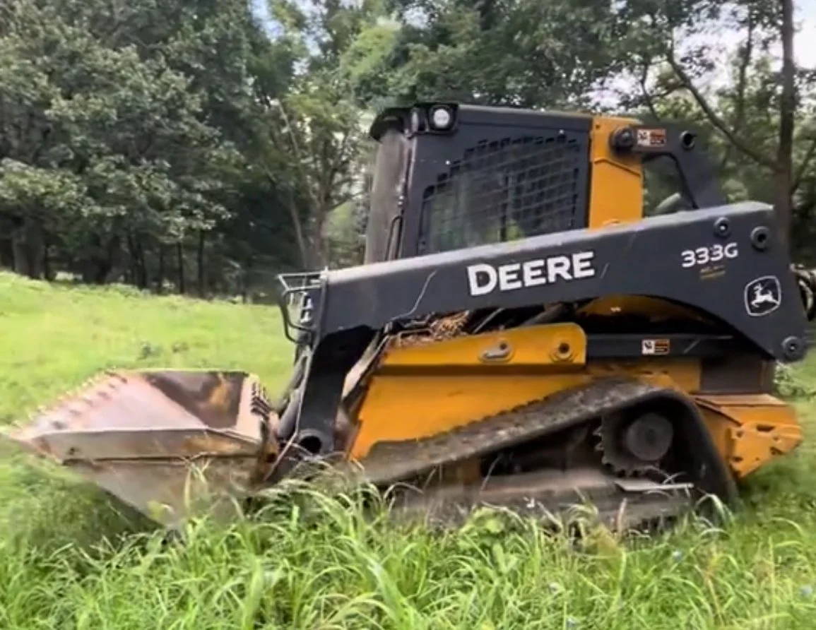 A yellow and black John Deere 333G compact track loader operating on a grassy area in a park or wooded area.