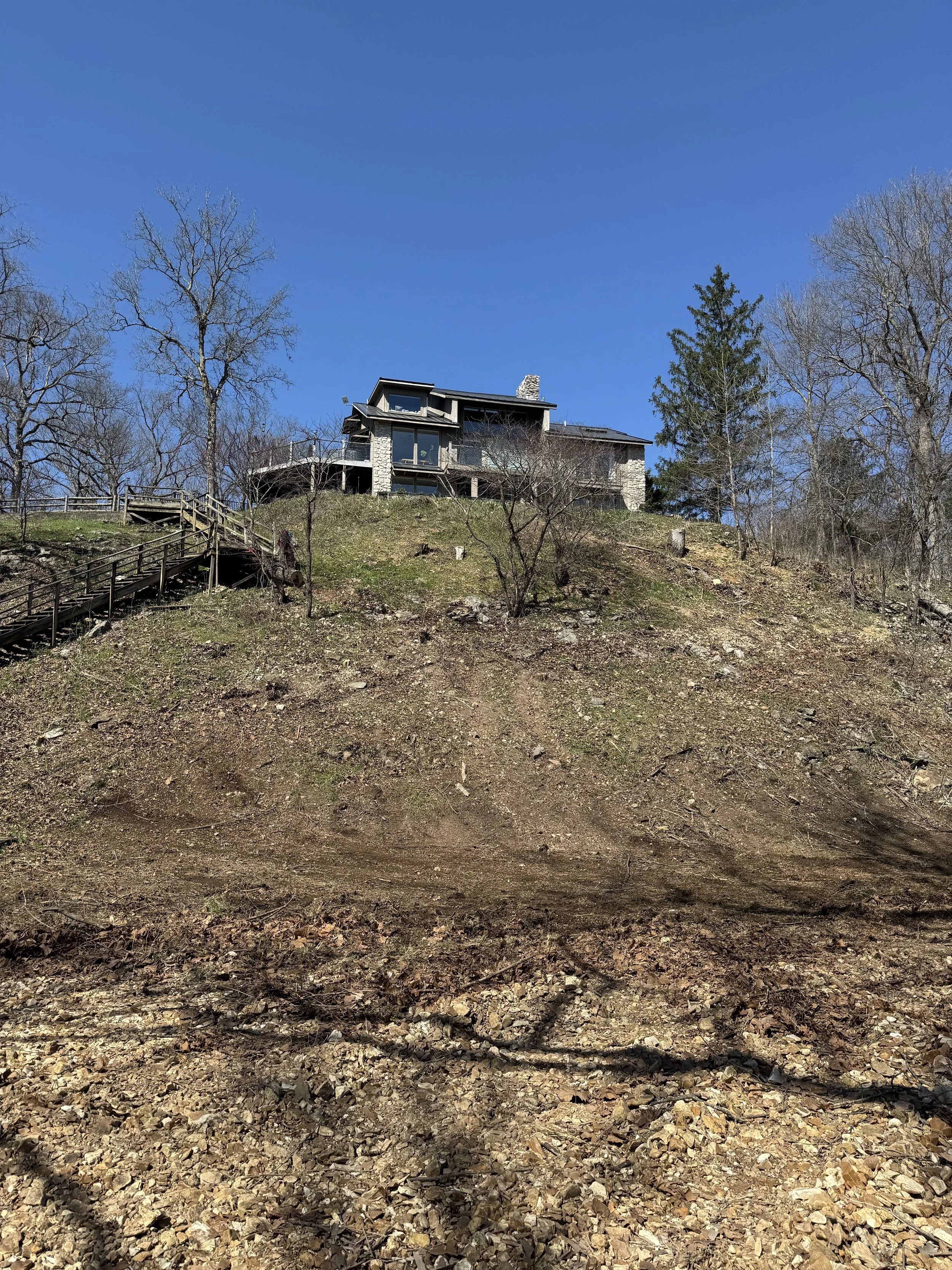 A house on a hill with a wooden staircase leading up to it, surrounded by leafless trees and a clear blue sky.