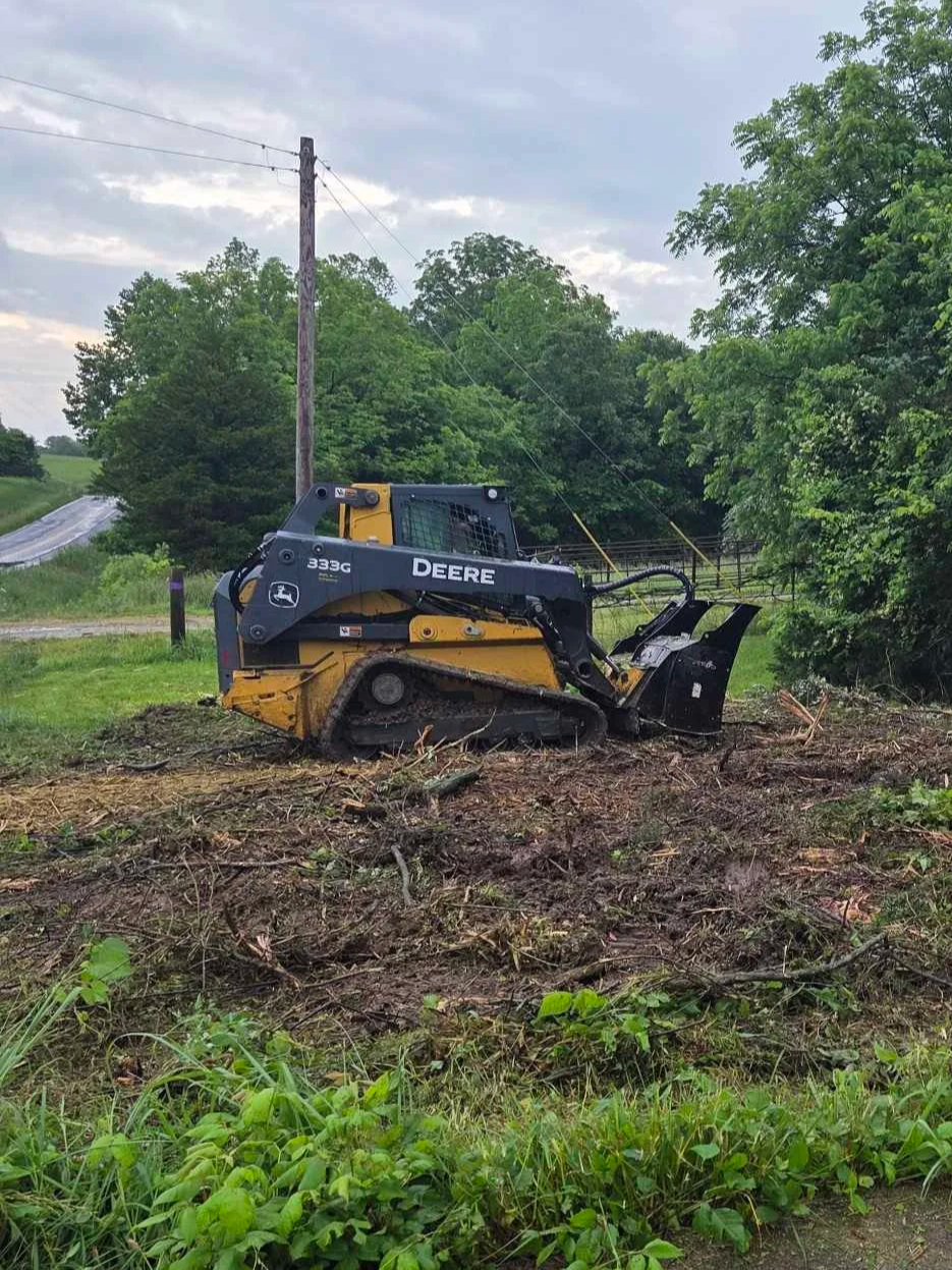 A small bulldozer working in a dirt clearing, surrounded by green trees and grass, with a partly cloudy sky overhead.