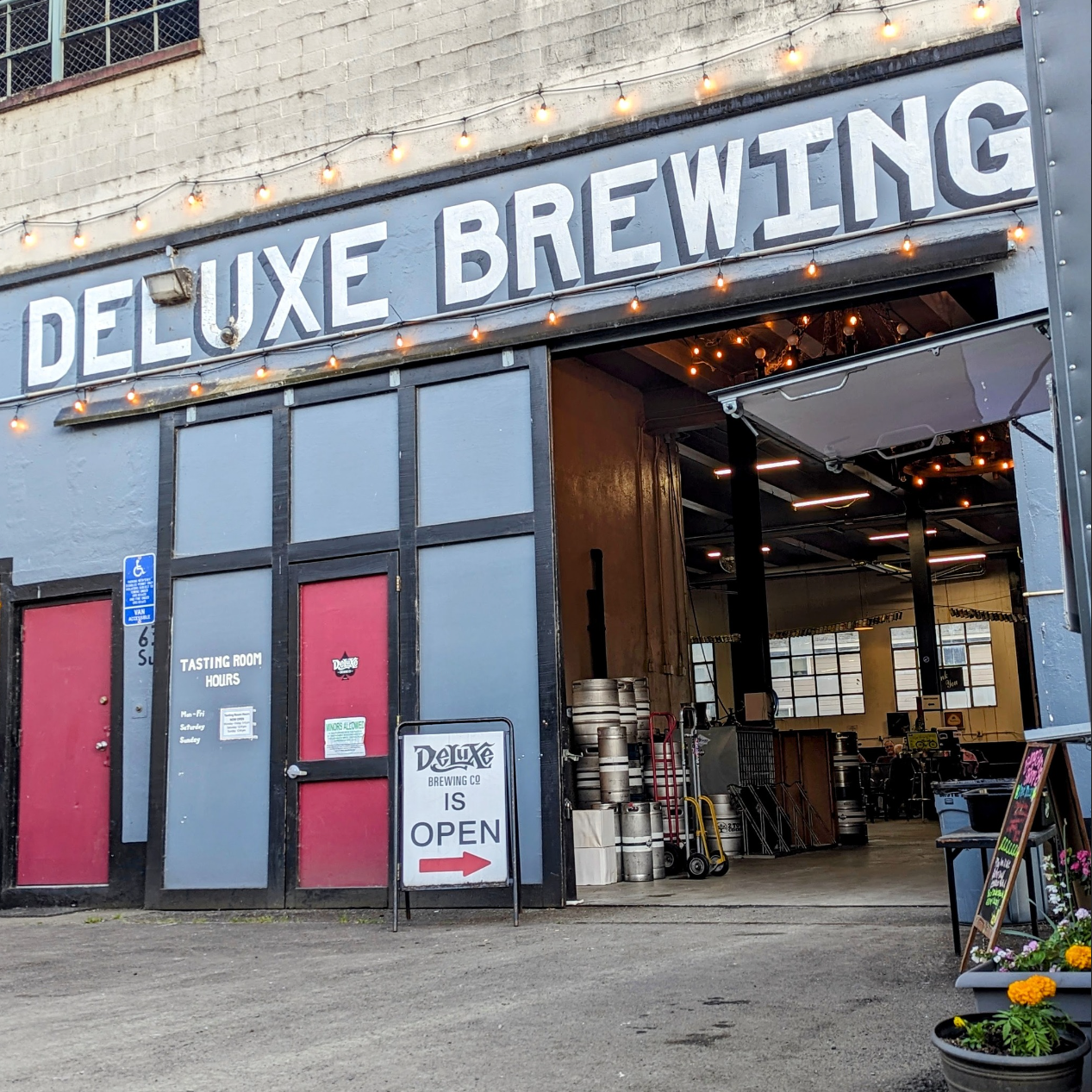 The exterior of Deluxe Brewing Co. with a large sign above the entrance, string lights, kegs stacked outside, and a sign indicating the brewery is open.