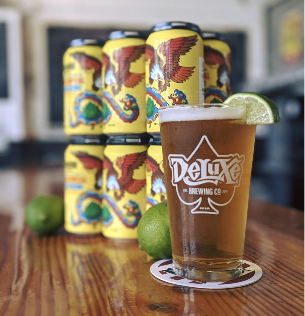 A glass of beer with a lime wedge on the rim, placed on a coaster on a wooden table, with several cans of beer with a dragon and eagle design in the background and two whole limes nearby.