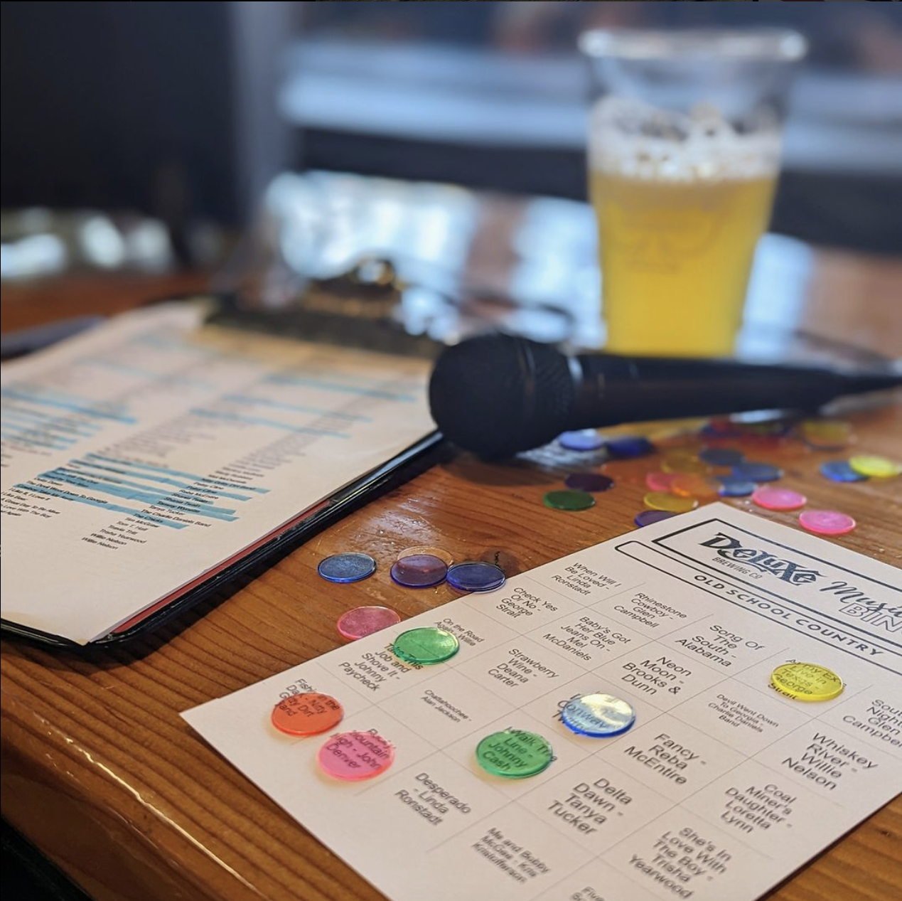 Music Bingo is on Thursday nights. A wooden table with a microphone, a sheet of paper with song titles, and music bingo markers scattered on it.