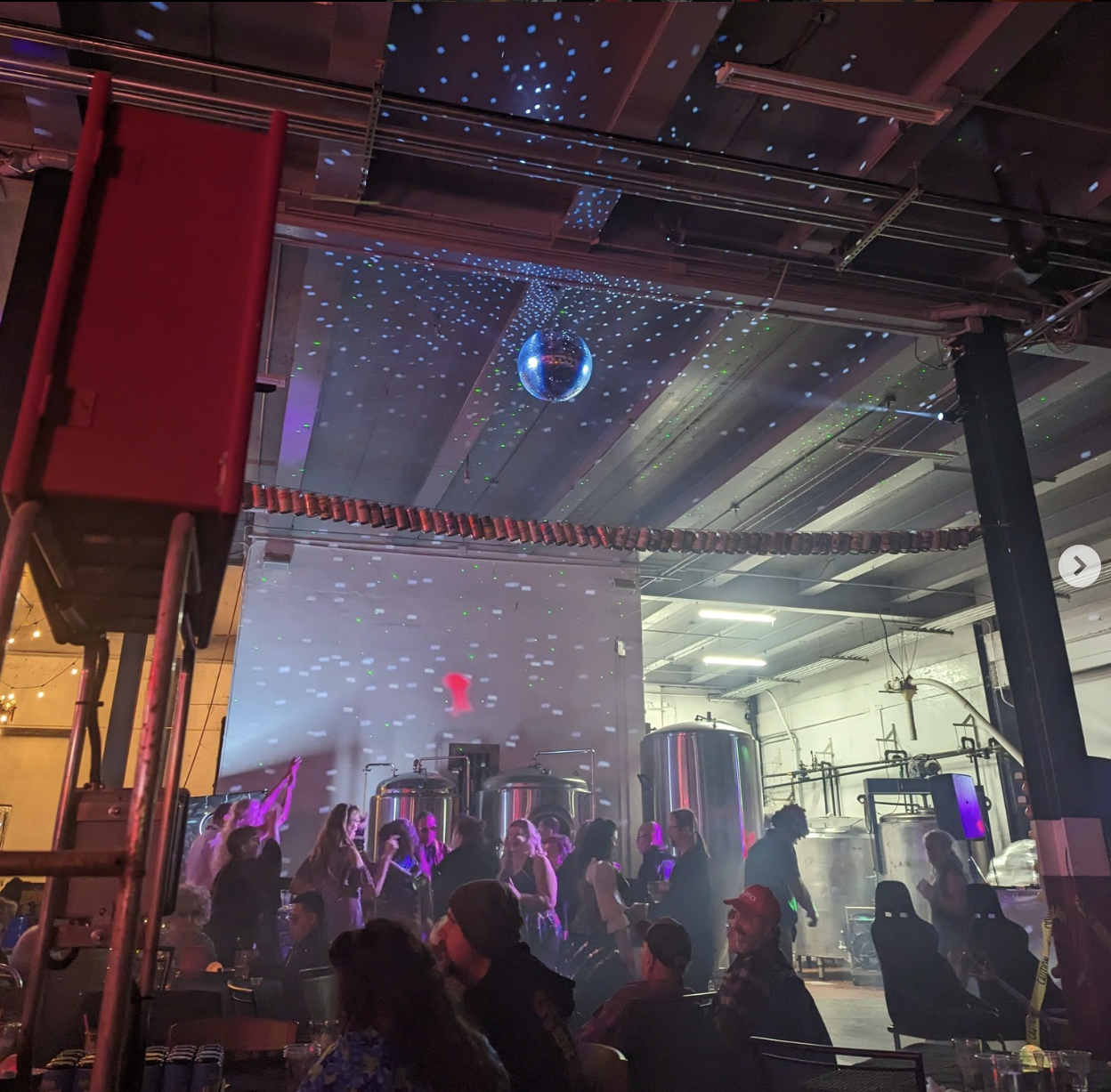 People dancing and socializing in a brewery, industrial-style venue with a disco ball and colorful lights.