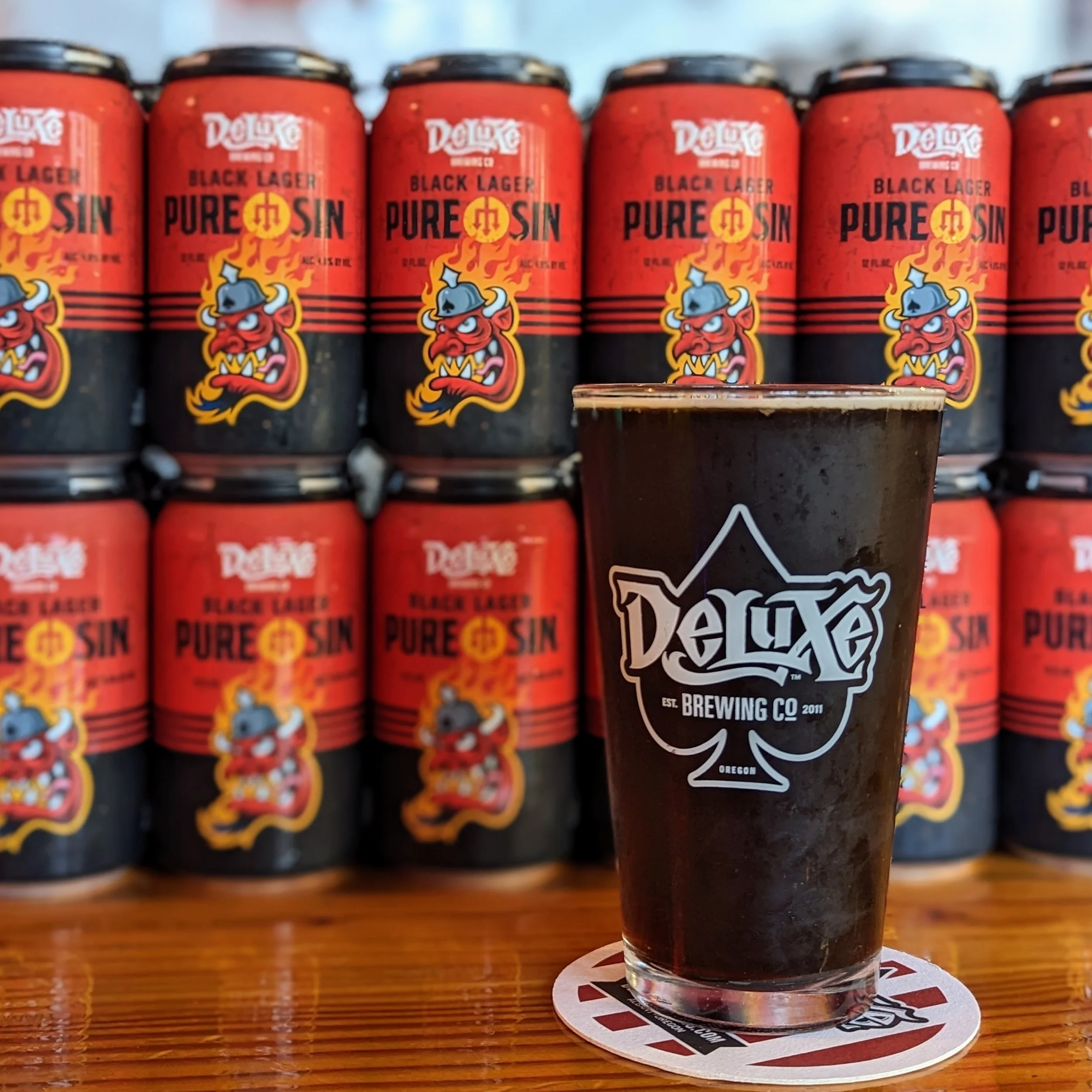 A pint glass of dark beer on a coaster with a label from Deluxe Brewing Company. In the background, there are several cans of Deluxe Black Lager beer with a red and black label.