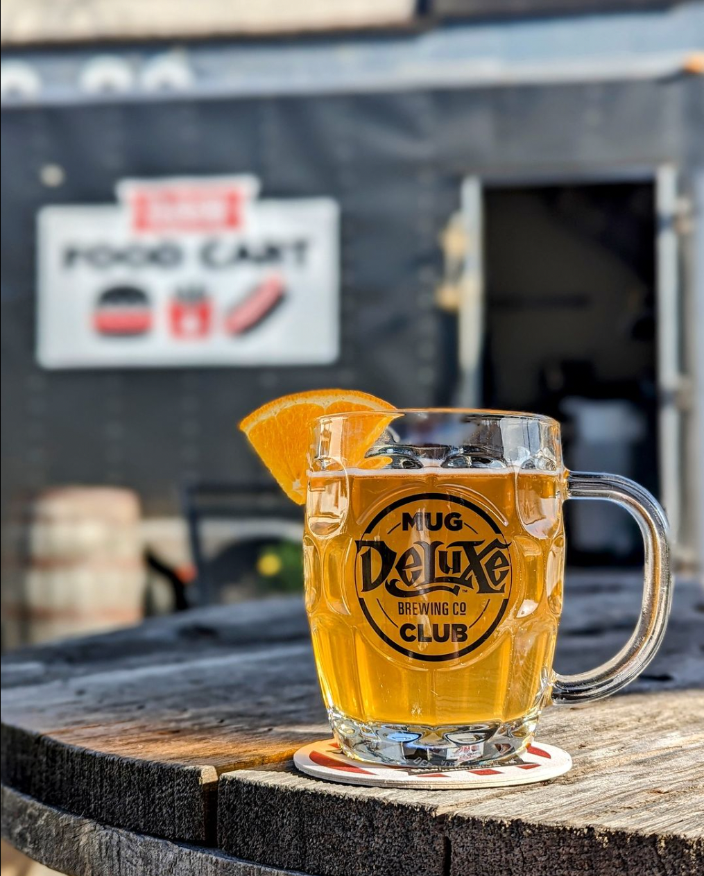 A glass mug with the logo 'Deluxe Brewing Mug Club' filled with a beer and garnished with an orange slice, placed on a wooden table outdoors.