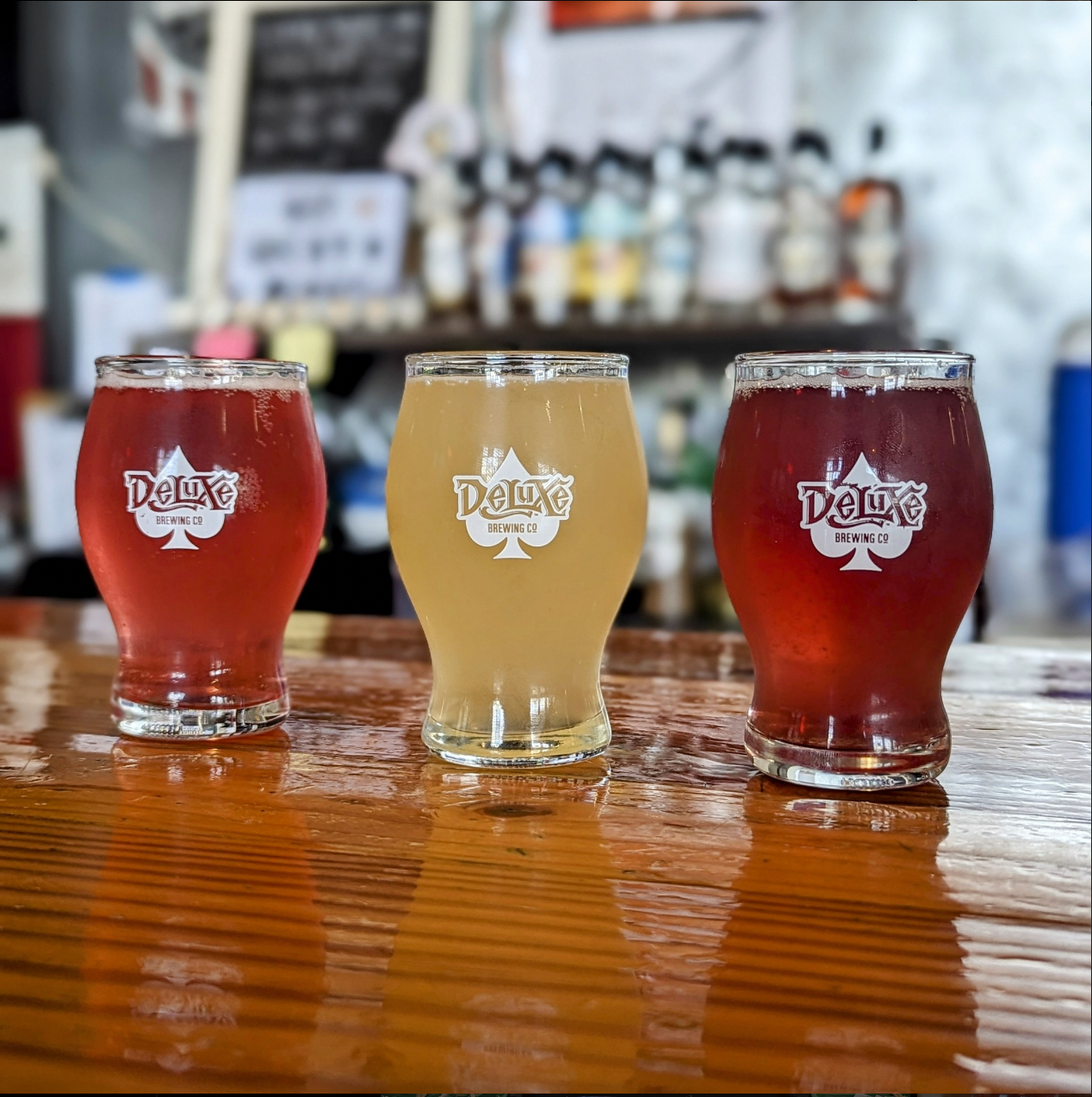 Three glasses of beer on a wooden bar, from left to right: Amber Lager, Pilsner, IPA. All glasses have the 'Deluxe Brewing Co' logo.