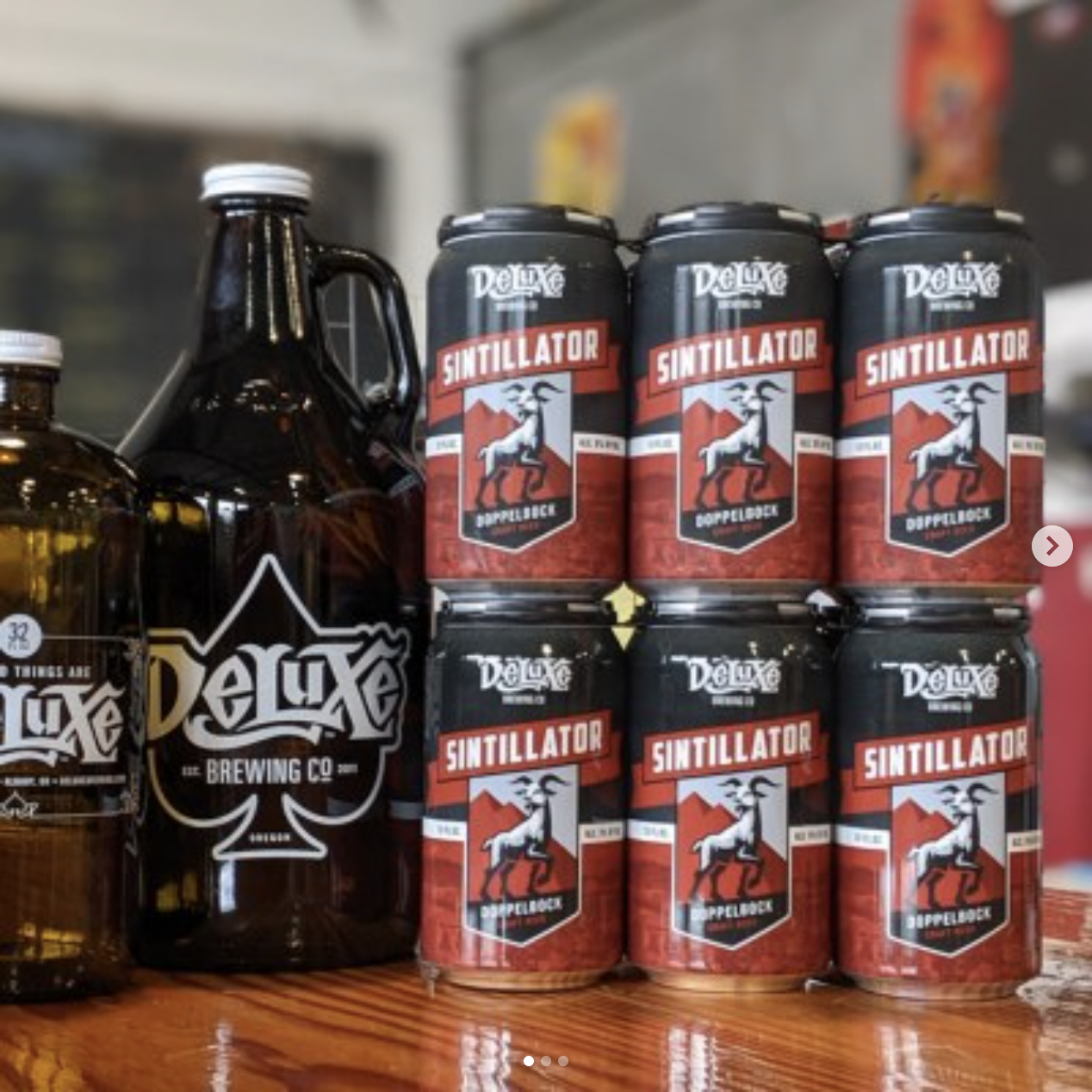 Six cans of Deluxe Sintillator beer on a wooden surface, with two bottles of Deluxe Brewing Co. beer to the left. The cans are black with red, white, and blue labels featuring a mountain goat and the words "Sintillator" and "Doppelbock."