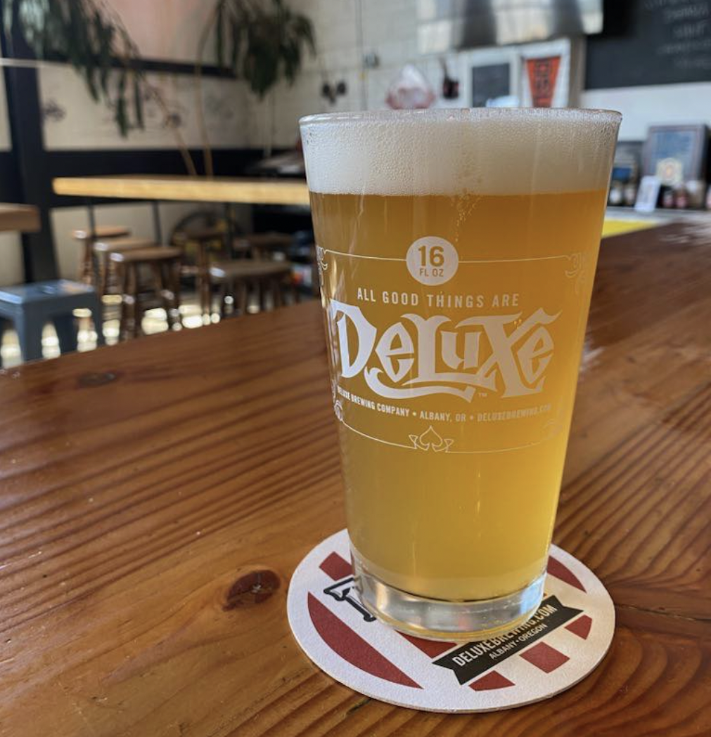 A glass of beer with a foamy head sitting on a round drink coaster on a wooden bar top inside the brewery.
