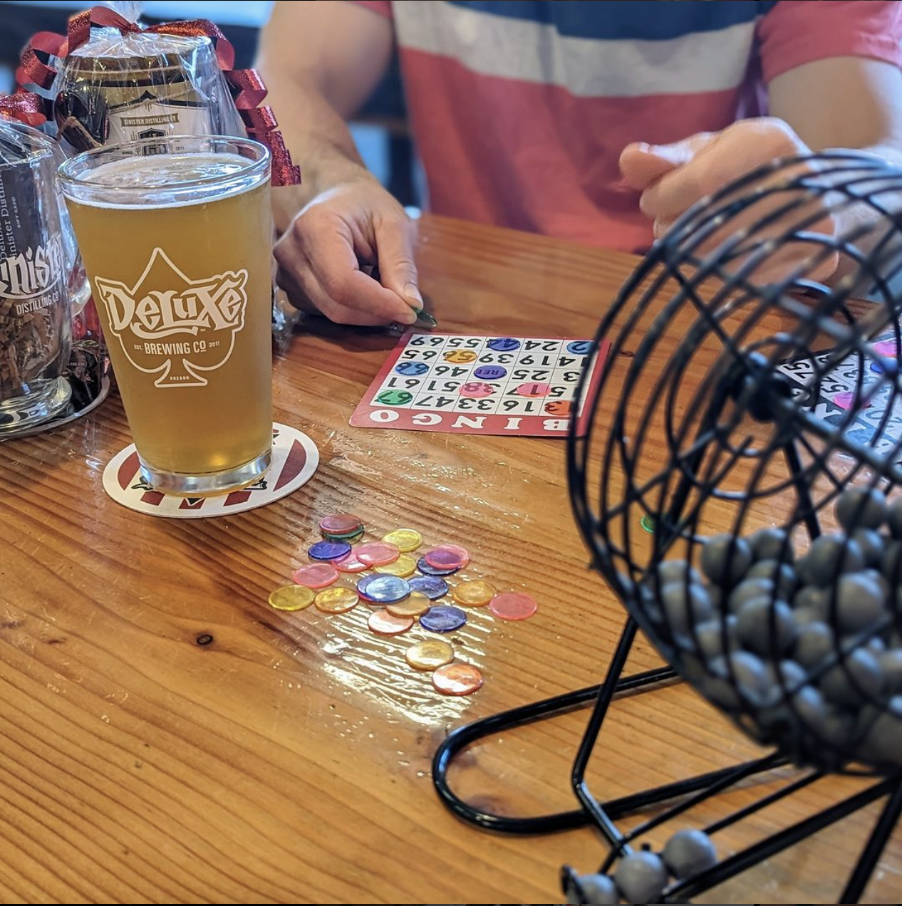 Two people playing bingo at a wooden table with a glass of beer, bingo cards, and colorful plastic bingo chips. One person is calling Bingo.