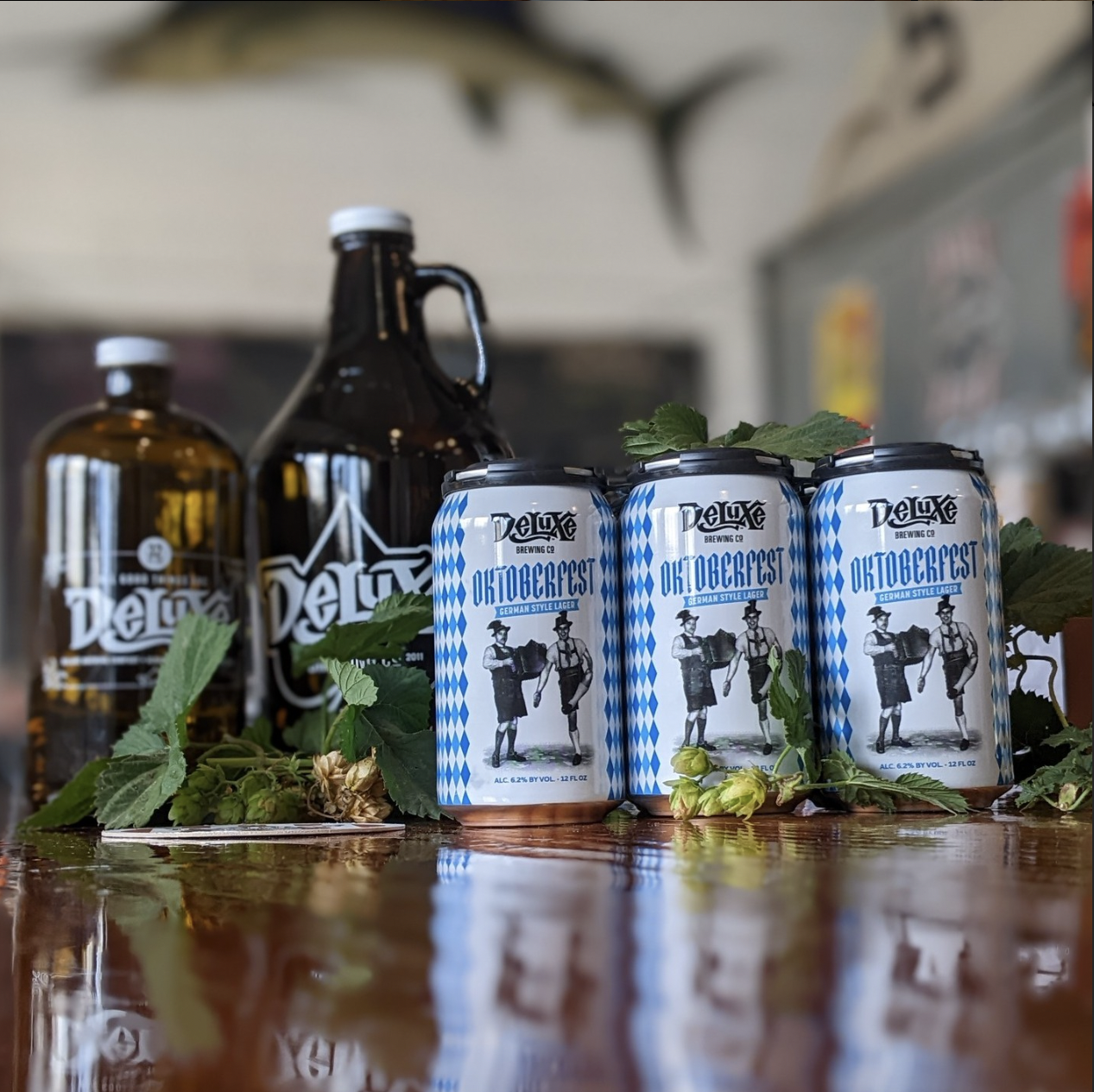 Three cans of Oktoberfest German-style lager beer on a wooden surface, surrounded by green hops leaves and flowers, with two brown glass bottles of DeLuxe beer and a blurred background.