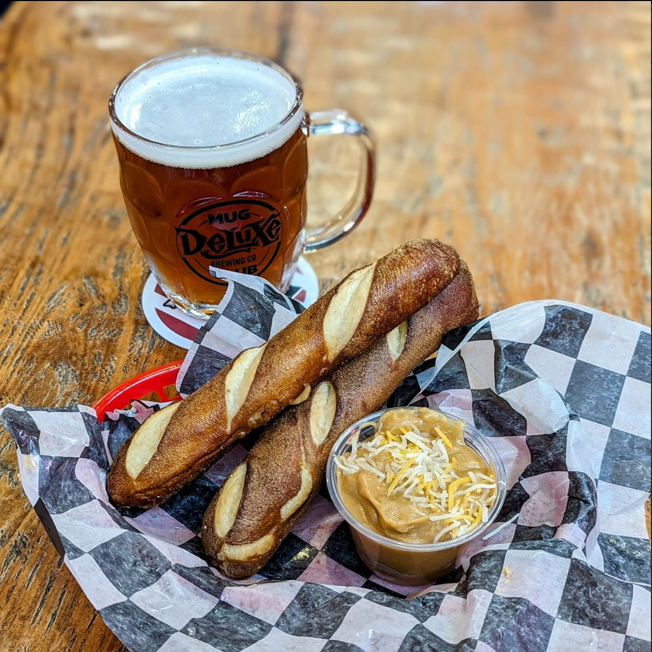 A basket with two pretzels and a small container of cheese dip topped with shredded cheese, accompanied by a mug of beer on a wooden table.