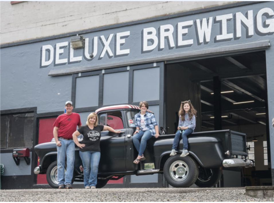 Four people, all family, posing near a vintage black truck outside a building with a sign that reads 'Deluxe Brewing'.