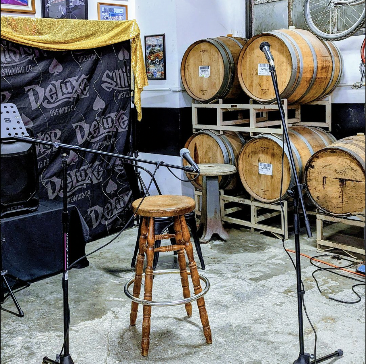 Stage area inside a brewery with barrels, microphones, stand-up stool, and dark background curtain with logo for Open Mic night.