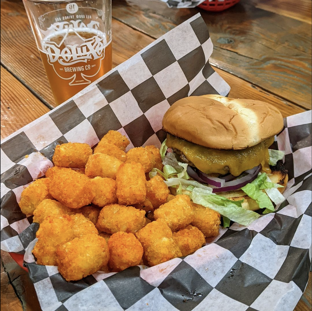Basket with tater tots and a cheeseburger with lettuce, tomato, onions, pickles, and melted cheese on a bun, alongside a glass of beer on a wooden table.