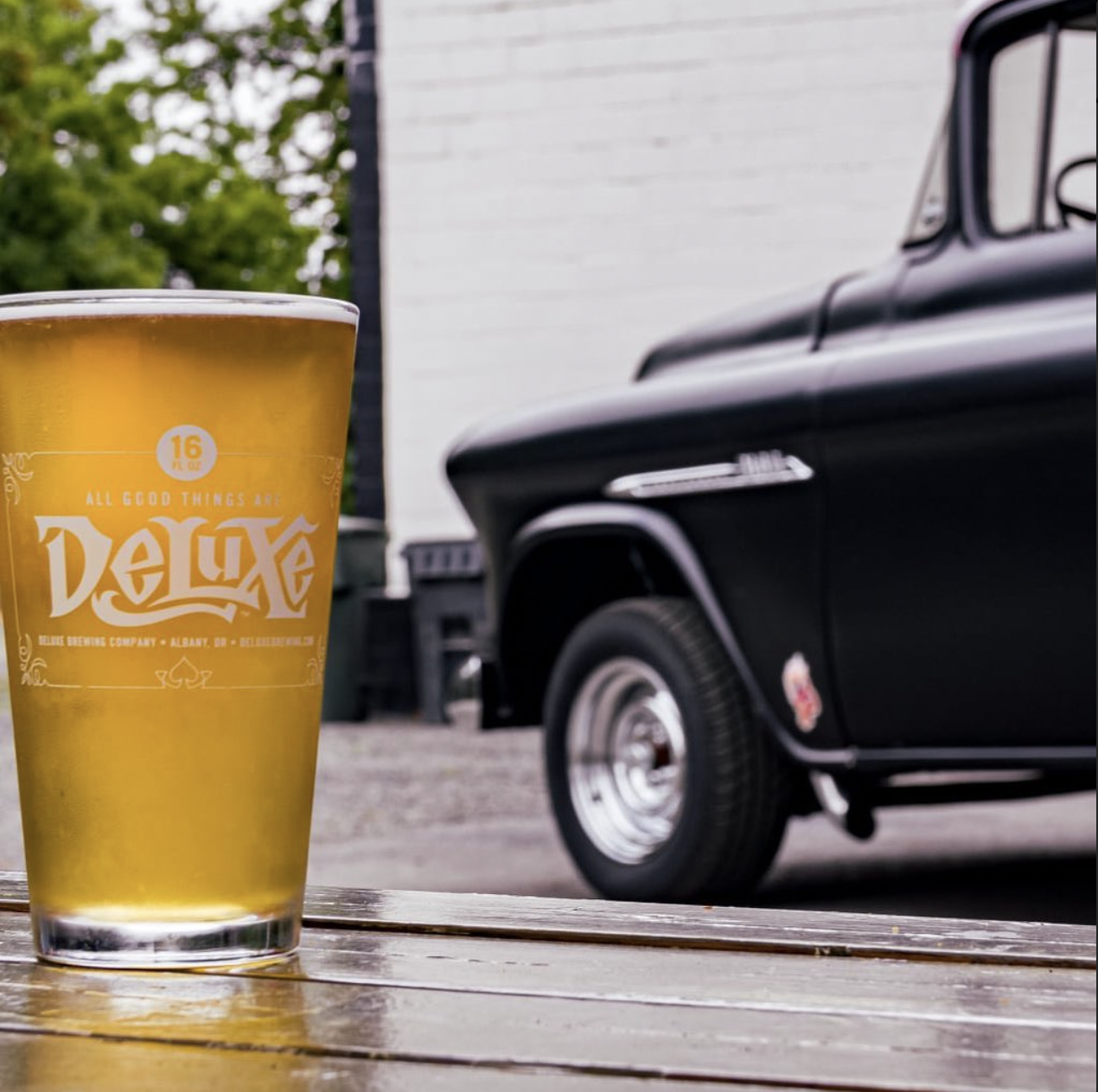 A glass of beer with a logo that reads 'Deluxe' on a wooden table outdoors, with a vintage black car and green trees in the background.