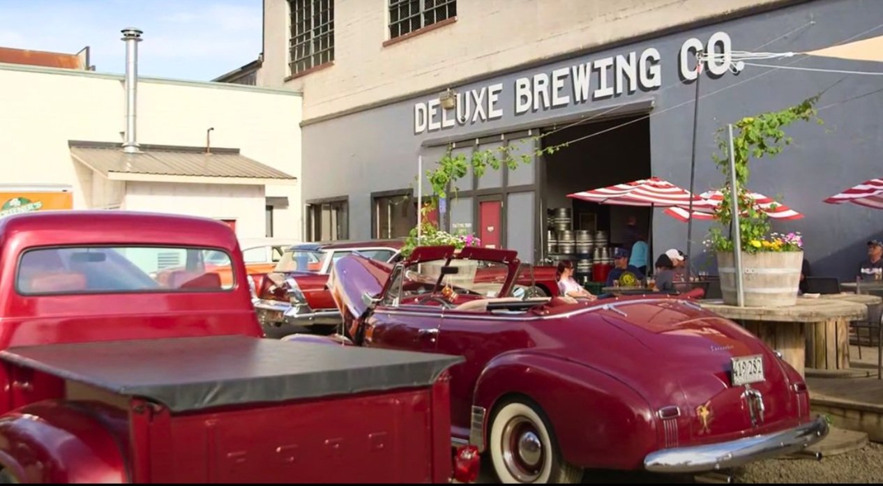 An outdoor scene in front of Deluxe Brewing Co featuring vintage cars, including a red convertible with its top down, parked on the street. There are people sitting under red and white striped umbrellas outside the brewery, which has a gray exterior with large windows and the sign "DELUXE BREWING CO" above the entrance.