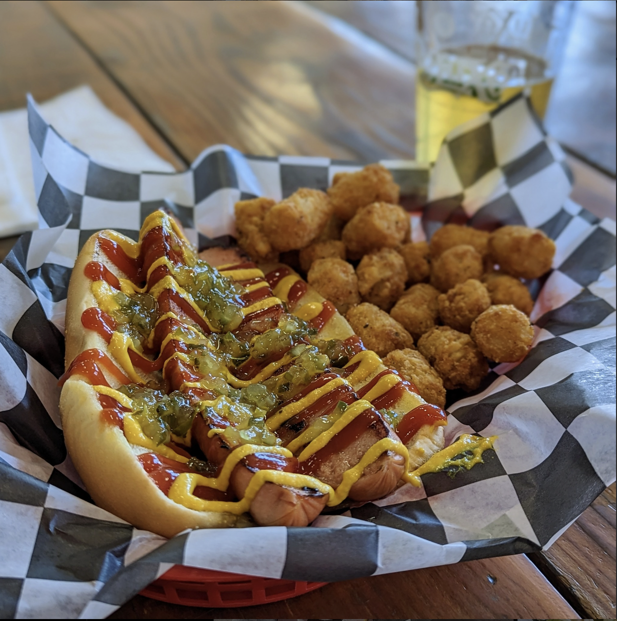 Hot dog topped with mustard, ketchup, relish, and mustard, served with fried tater tots and a glass of beer on a checkered paper-lined basket.