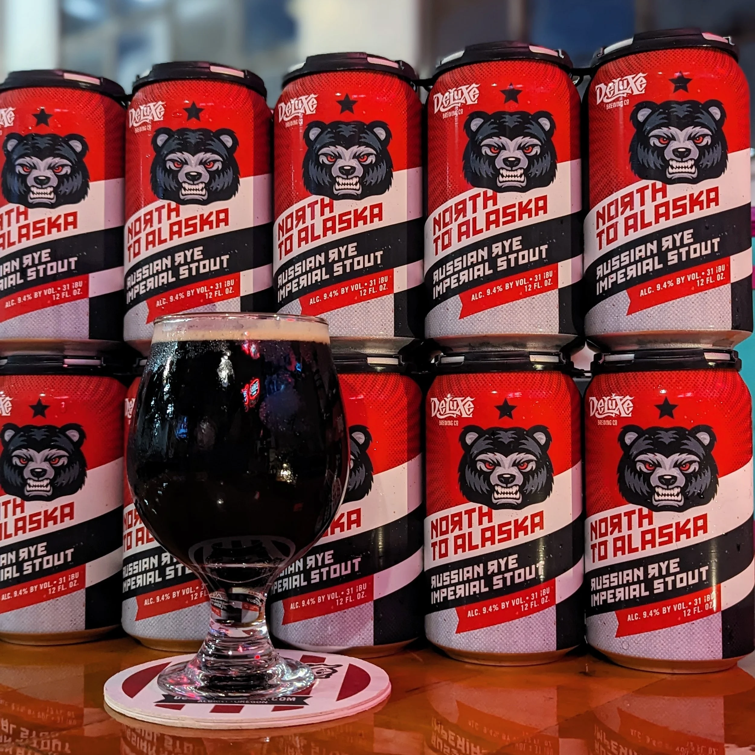 Six cans of North to Alaska Russian Style Imperial Stout beer with a black bear graphic, red and white design, and alcohol content of 9.4%, lined up on a shelf. A glass of dark stout beer placed in front of the cans, on a coaster and a wooden table.