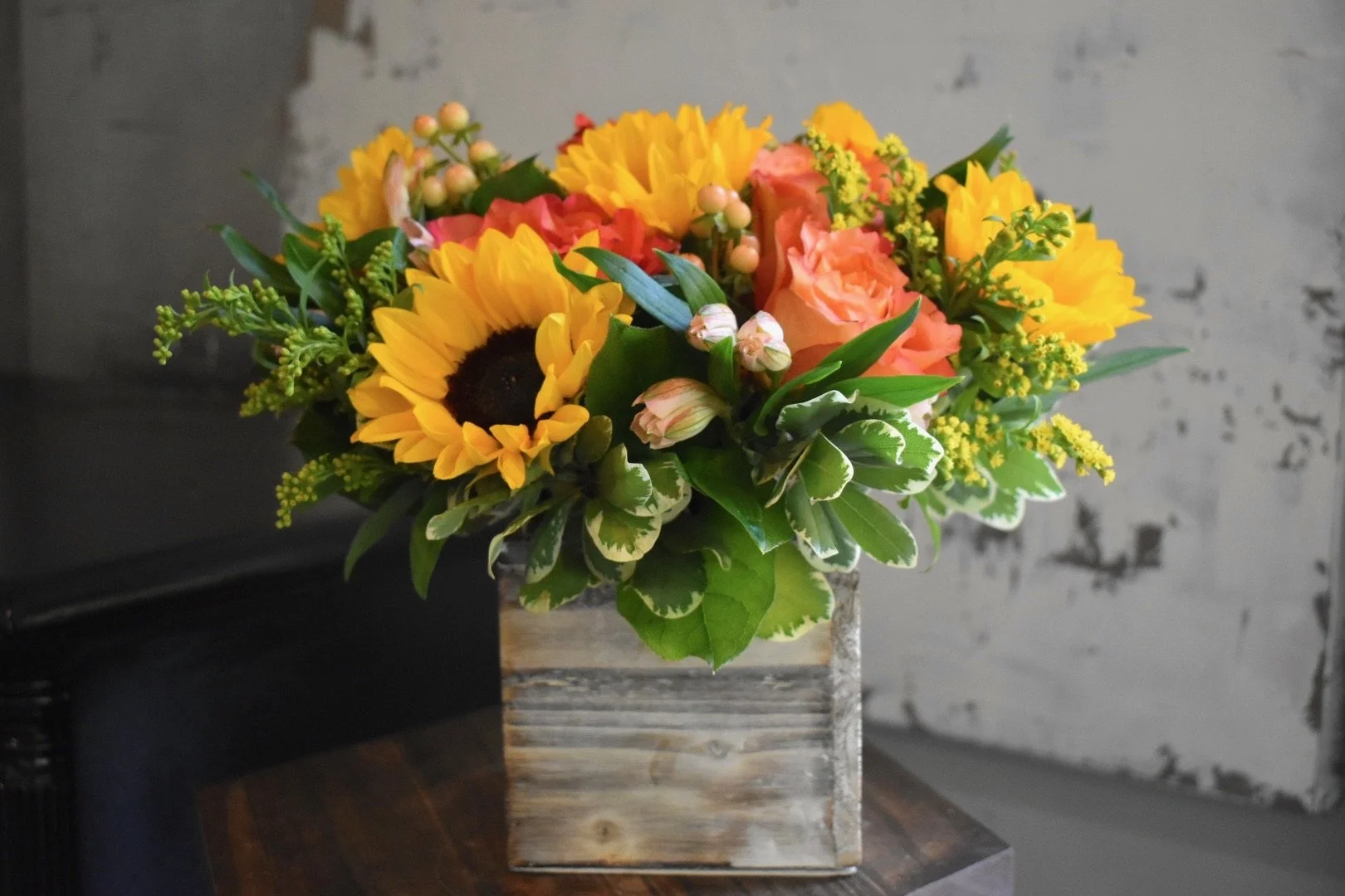 A bouquet of colorful flowers including sunflowers, pink roses, and variegated leaves in a rustic wooden box on a dark wooden surface.