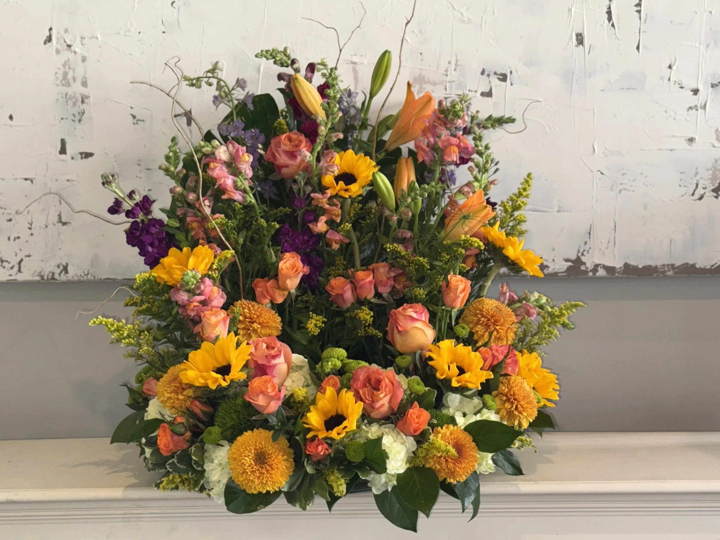 Colorful mixed flower arrangement featuring sunflowers, lilies, roses, and other blooms in shades of yellow, pink, purple, and white.