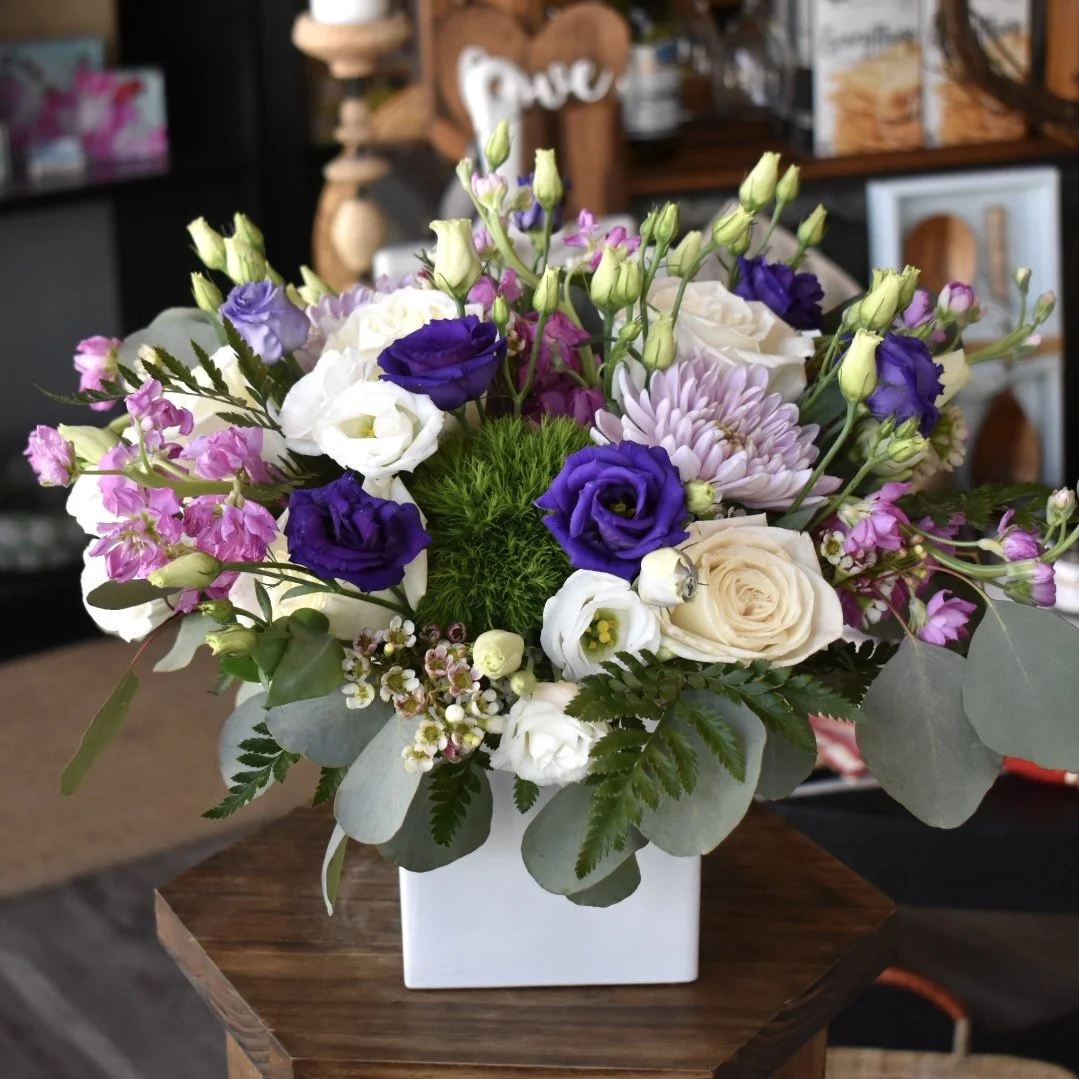A floral arrangement in a white square vase with purple, white, pink, and green flowers, placed on a wooden surface.
