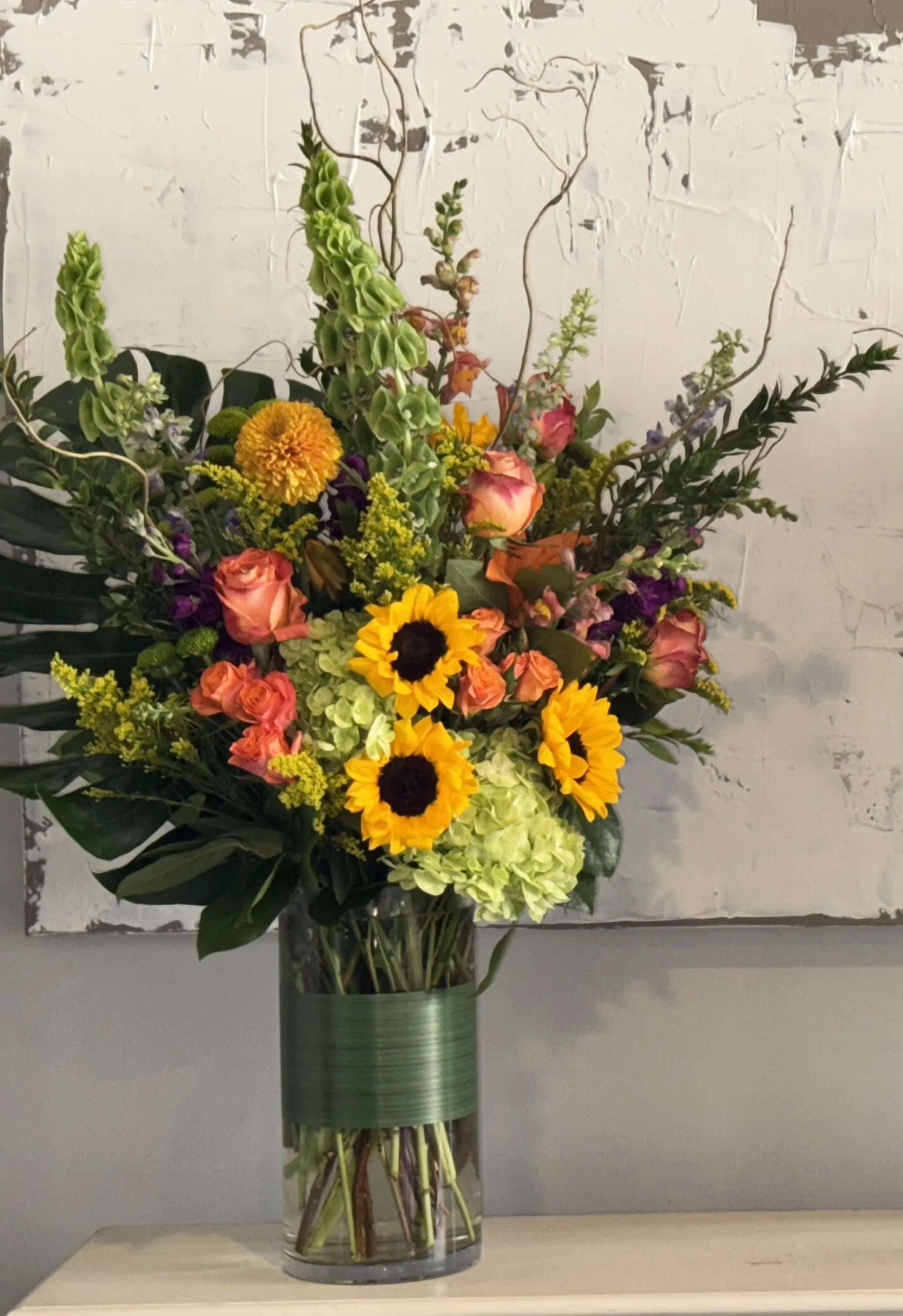 Colorful flower arrangement in a glass vase with sunflowers, roses, hydrangeas, and various greenery against a textured white wall.