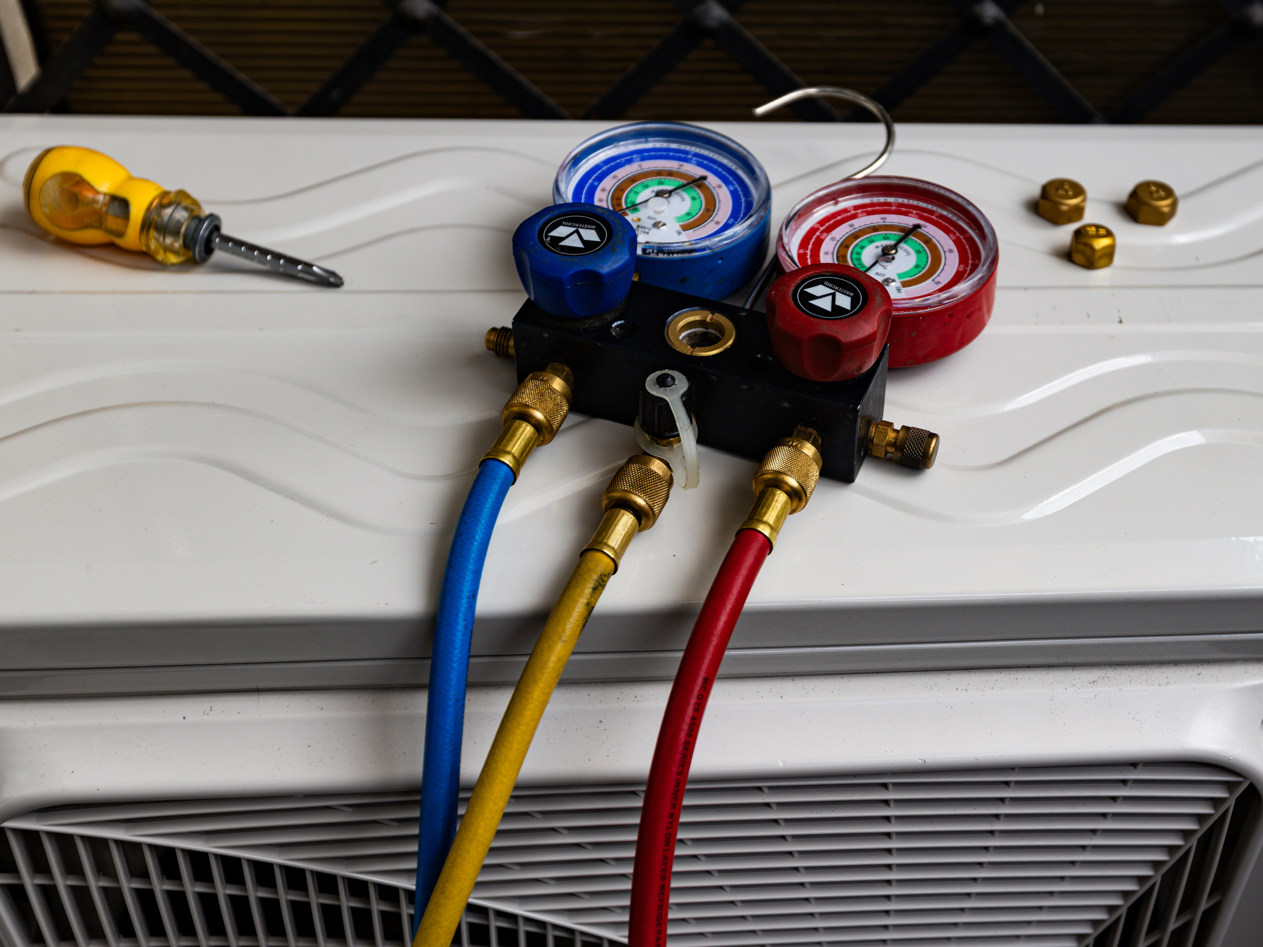 Air conditioning unit with manifold gauges and hoses attached for servicing, placed on top of a white surface.