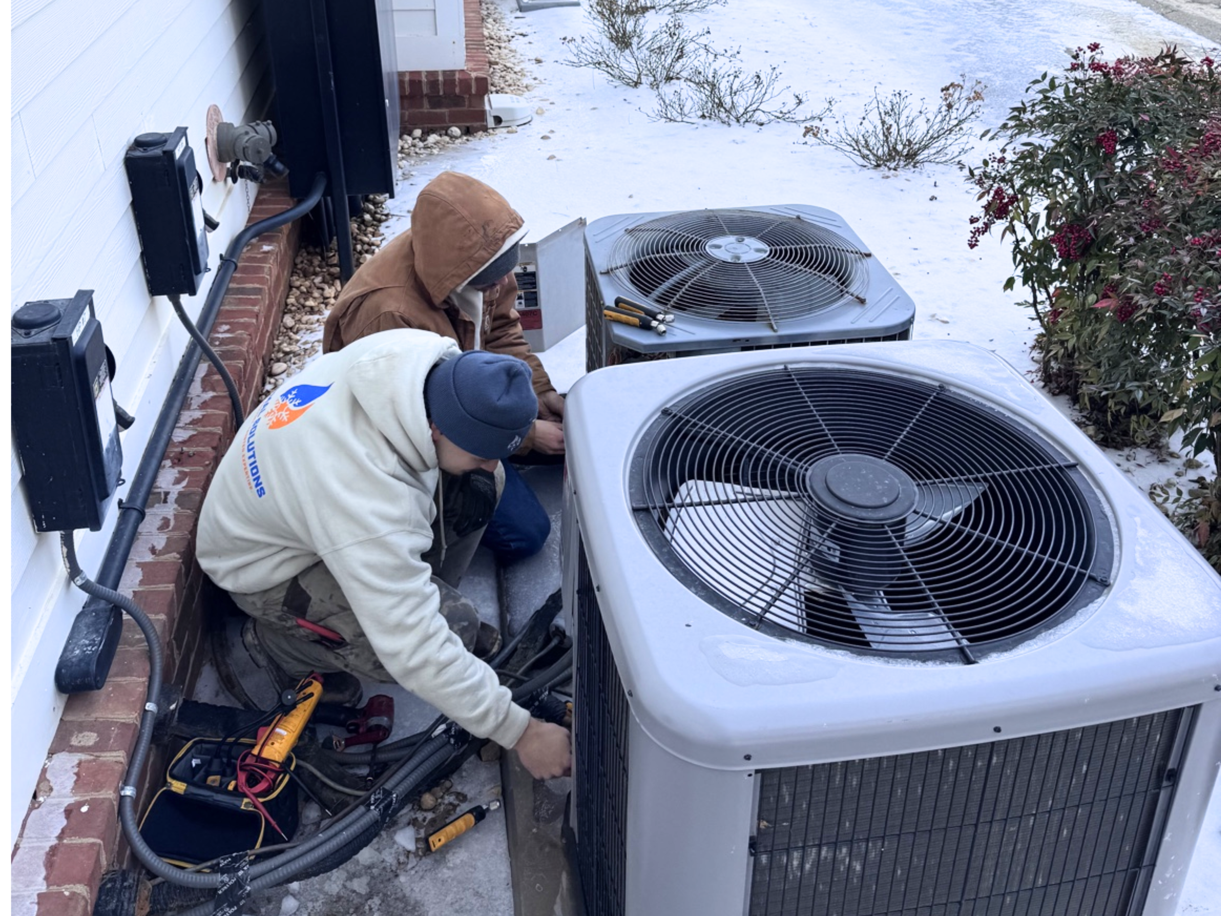 Two workers repairing or installing an air conditioning unit outside on a snow-covered ground beside a white house with brick foundation. One worker is wearing a tan hooded jacket, and the other is in a white hoodie with orange and blue logos. Tools and electrical equipment are scattered around.