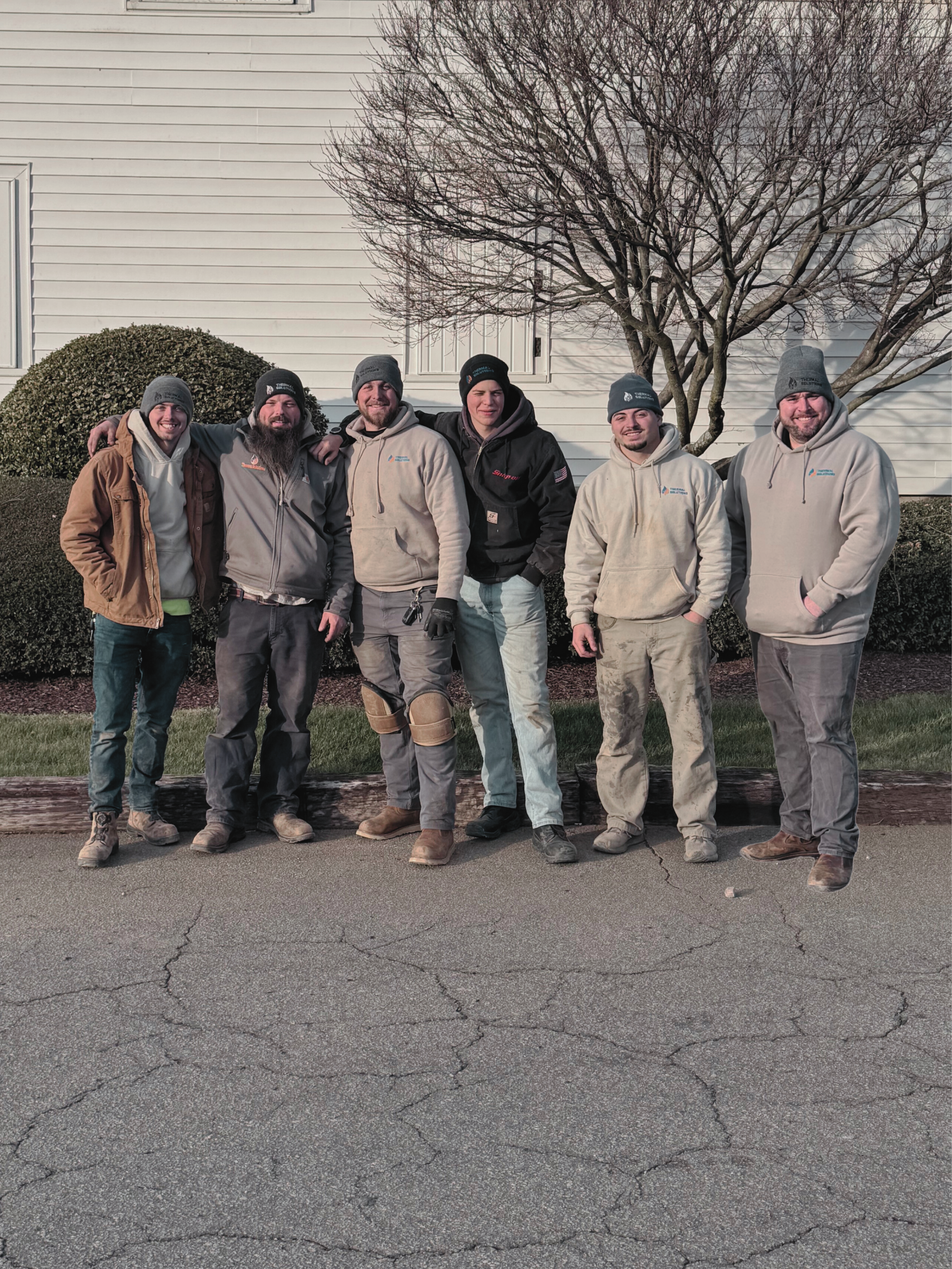 Group of six men standing outdoors in front of a white building with a leafless tree, all wearing winter clothing.