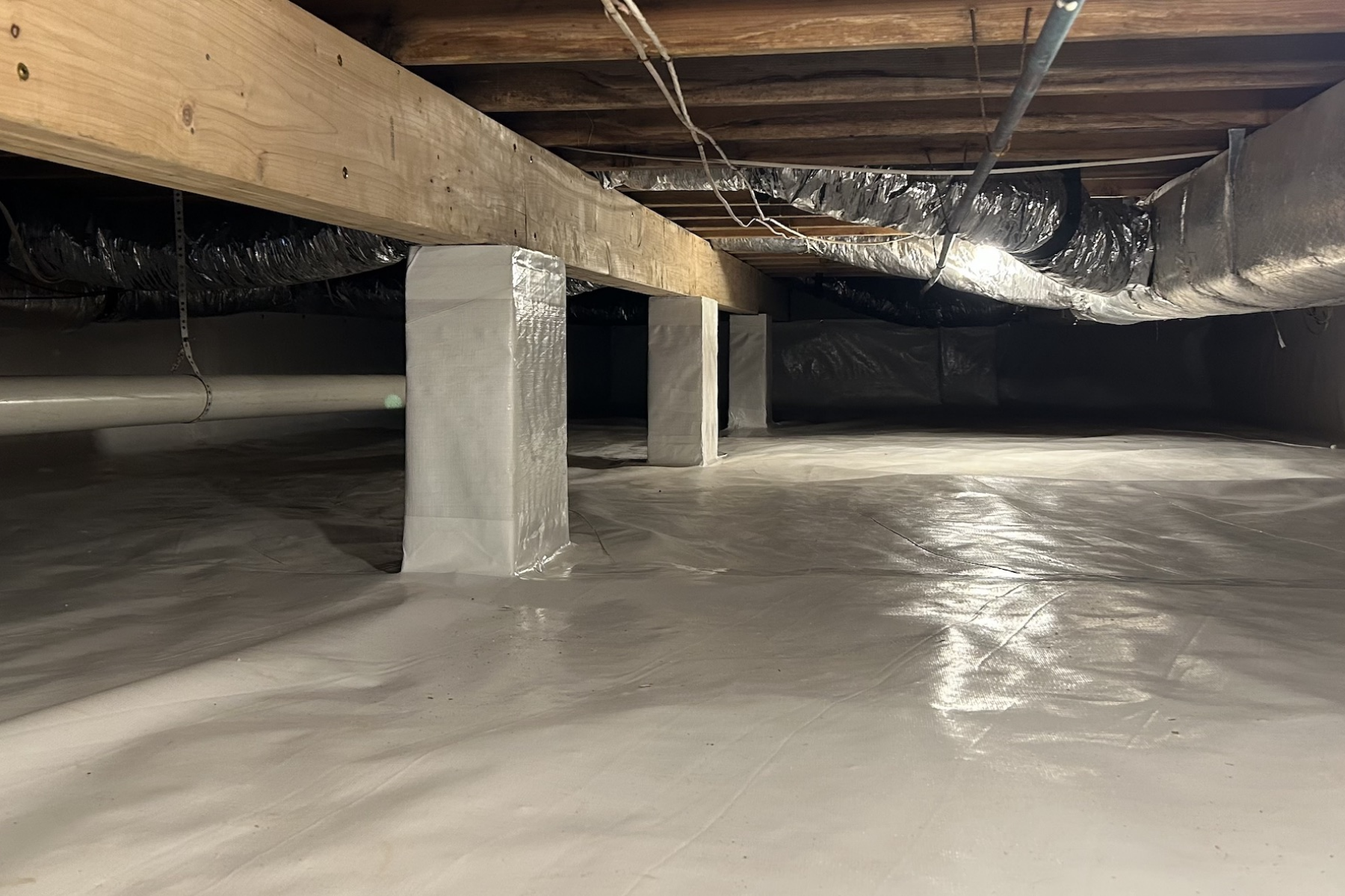 View of an unfinished basement or crawl space with insulated floor, wooden beams, and ductwork, supported by brick columns wrapped in insulation.